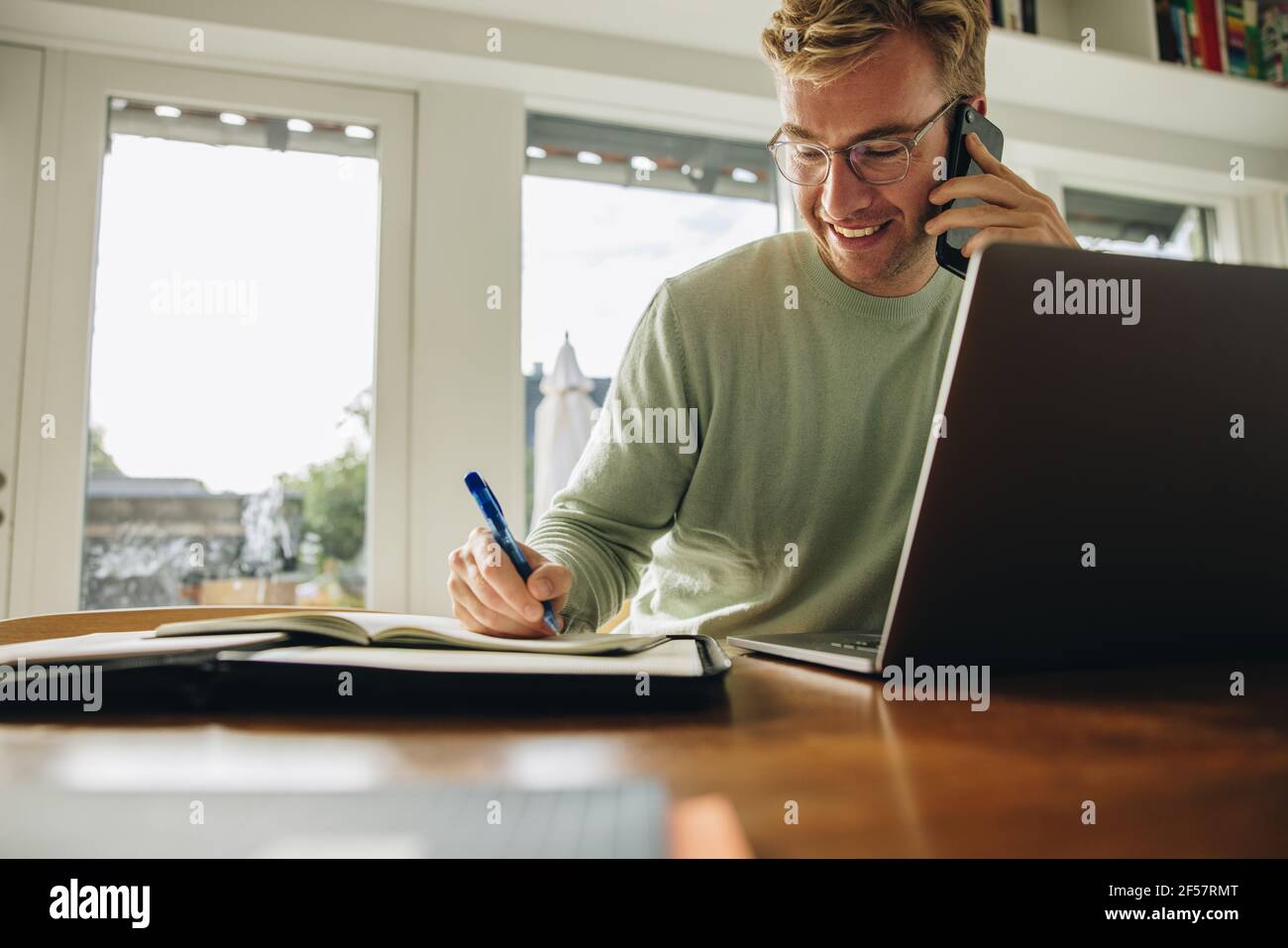 Uomo seduto al tavolo con un computer portatile che parla al telefono e scrive in un libro. Uomo che lavora da casa. Foto Stock