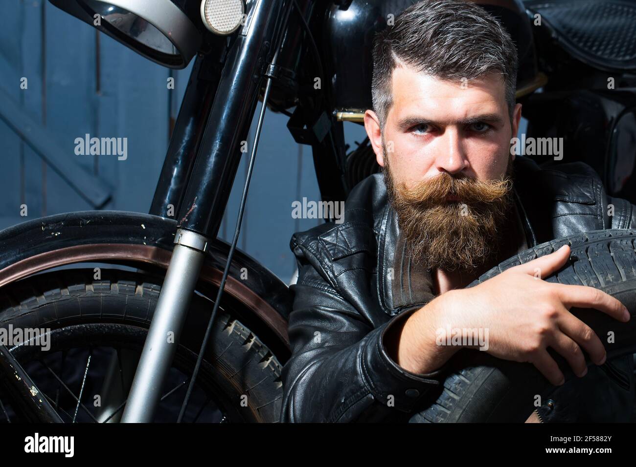 Bello bearded biker in giacca di pelle seduta vicino moto dentro garage Foto Stock