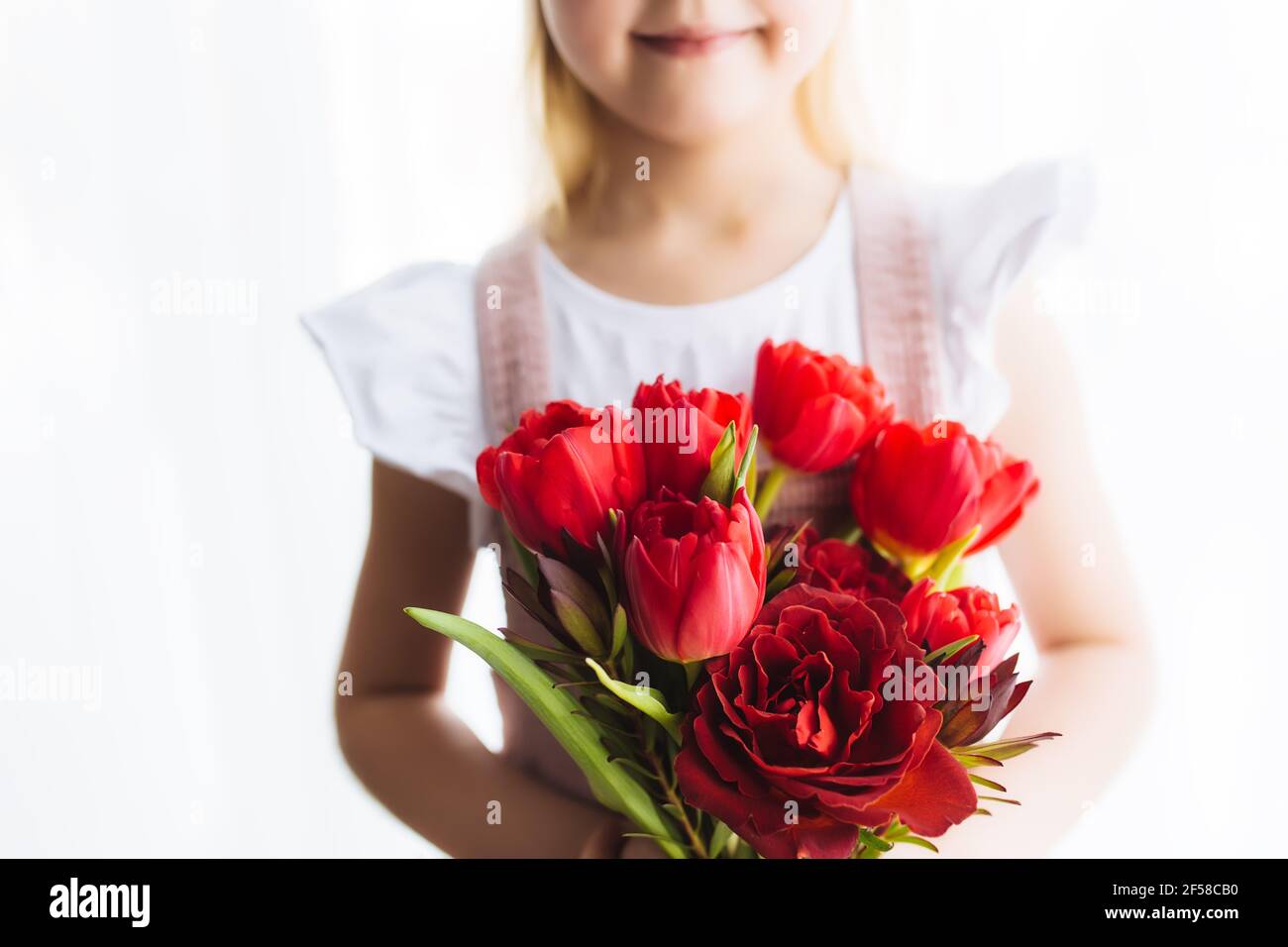 Piccola ragazza sorridente che tiene bouquet di fiori rossi di tulipano. Concetto di biglietto d'auguri per Pasqua, Festa della Madre, Giornata Internazionale della Donna, San Valentin Foto Stock