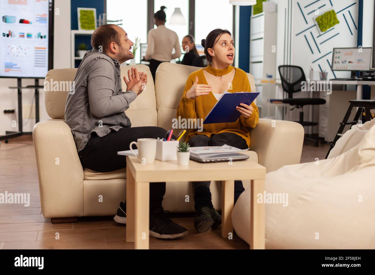 Uomo manager arrabbiato che sostiene una donna dipendente per un cattivo risultato di lavoro, seduto sul divano, mentre diversi colleghi che lavorano spaventato in background. Dipendente donna di affari che infuria sul lavoro difficile multitasking. Foto Stock