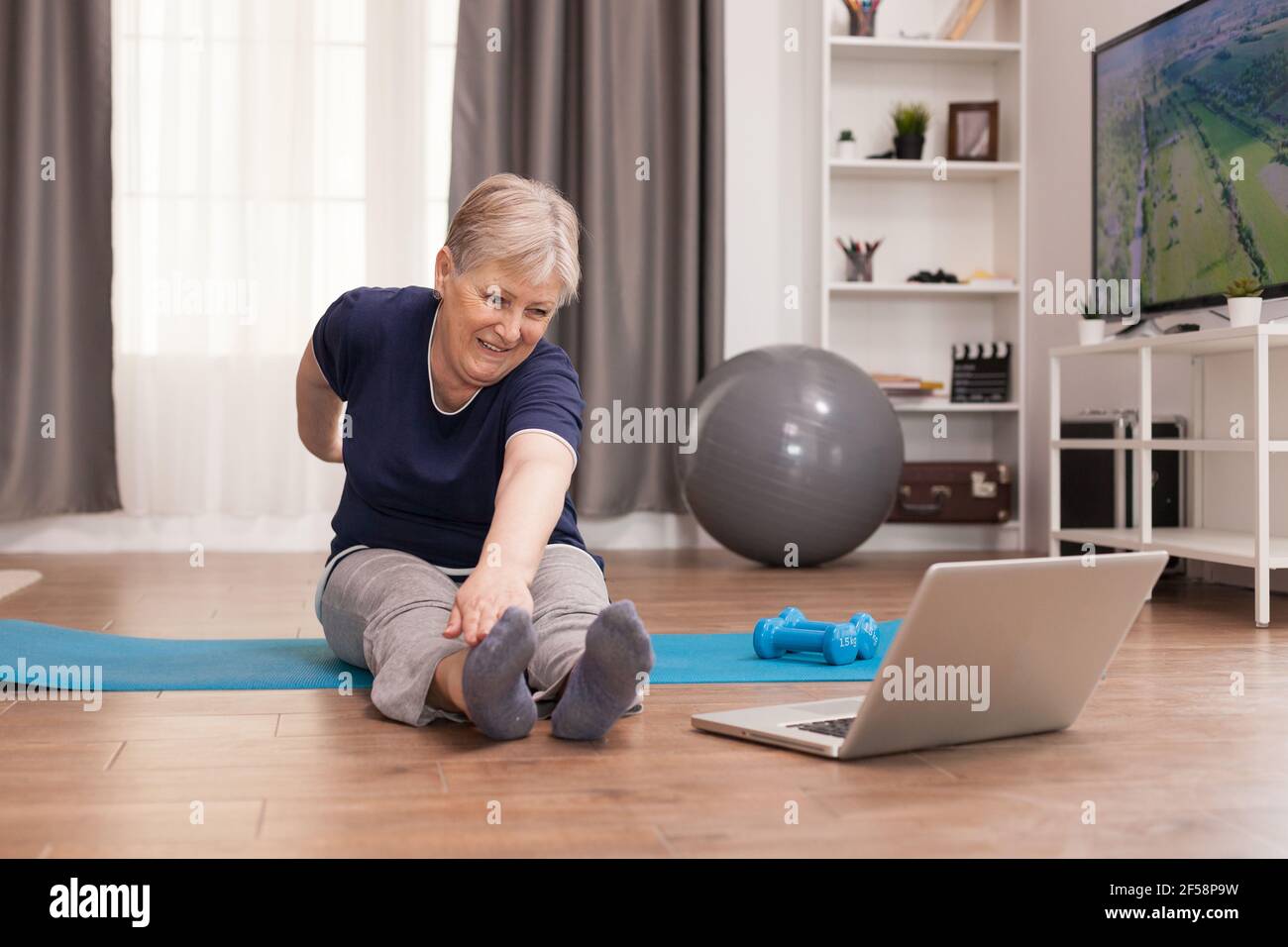 Allegra vecchia donna che fa sport guardando le lezioni online. Anziano pensionato online internet esercizio di formazione a casa attività sportiva con manubri, banda di resistenza, palla svizzera all'età di anziano pensionamento Foto Stock