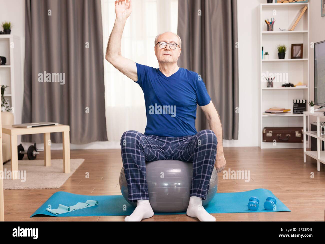 Pensionato che fa esercizio a casa con la palla svizzera. Anziano pensionato online internet esercizio di formazione a casa attività sportiva con manubri, banda di resistenza, palla svizzera all'età di anziano pensionamento. Foto Stock