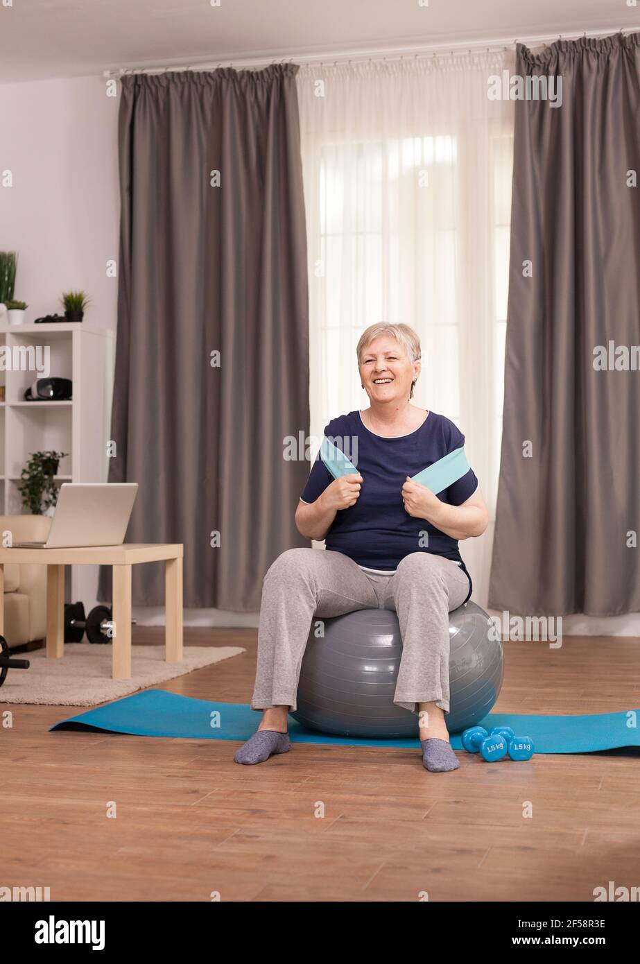 Donna anziana sana che usa la banda di resistenza per la vitalità. Anziano pensionato online internet esercizio di formazione a casa attività sportiva con dumbbell, palla svizzera in età pensionabile anziana Foto Stock
