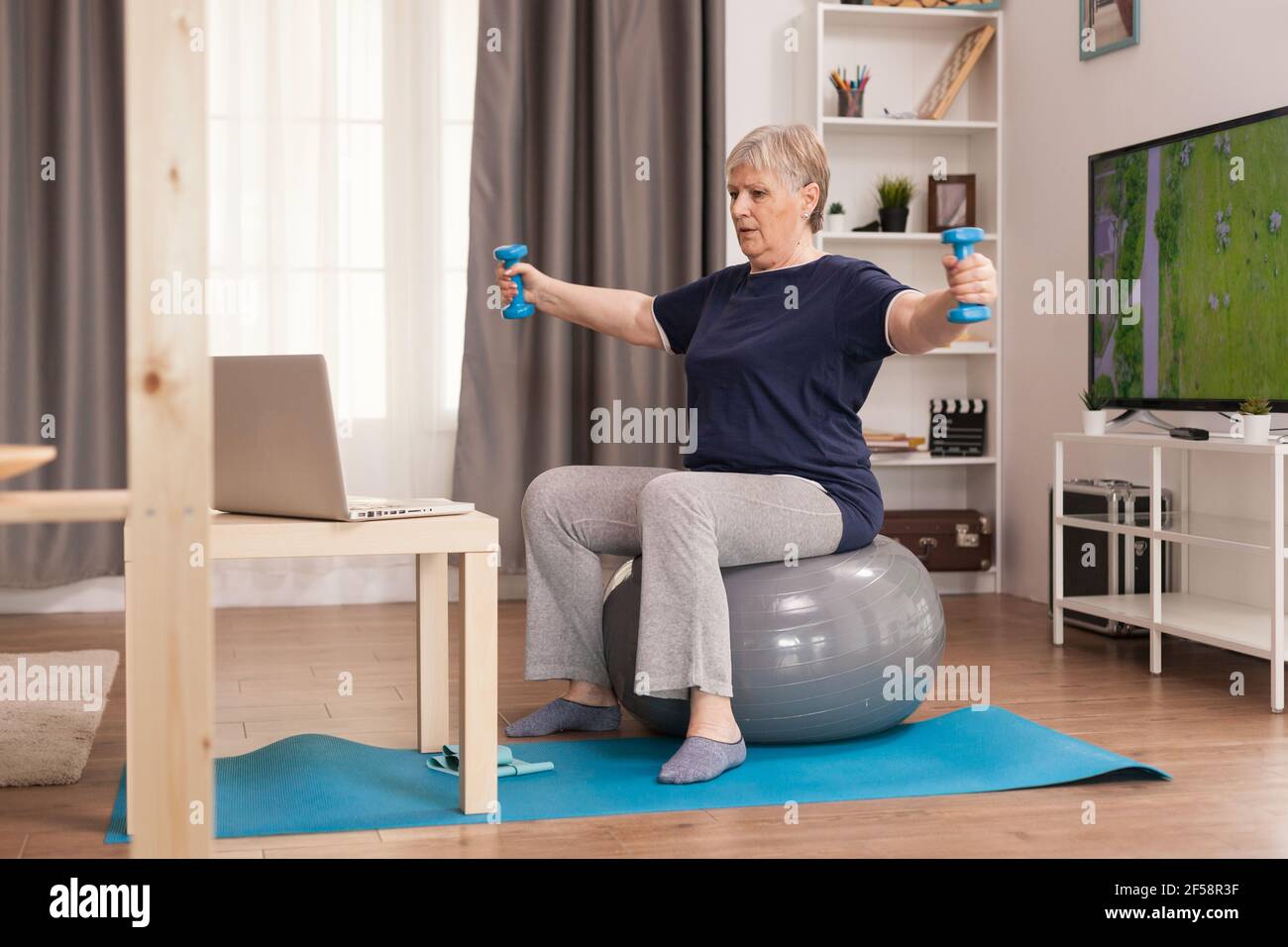 Donna ritirata allenando spalle con pesi. Anziano pensionato online internet esercizio di formazione a casa attività sportiva con manubri, banda di resistenza, palla svizzera all'età di anziano pensionamento Foto Stock