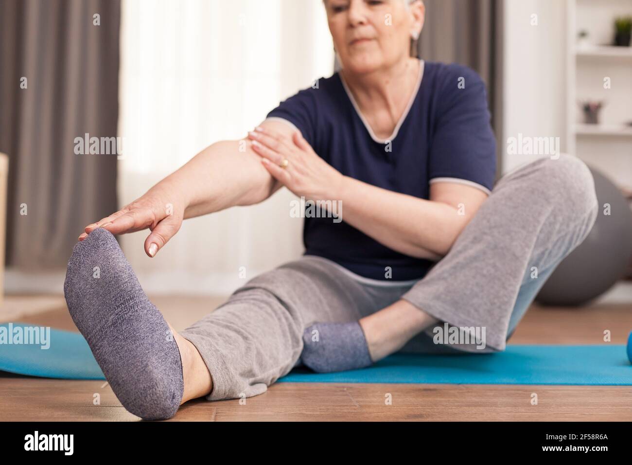 Nonna moderna che fa stretching a casa oon yoga tappetino. Anziano pensionato online internet esercizio di formazione a casa attività sportiva con manubri, banda di resistenza, palla svizzera all'età di anziano pensionamento Foto Stock