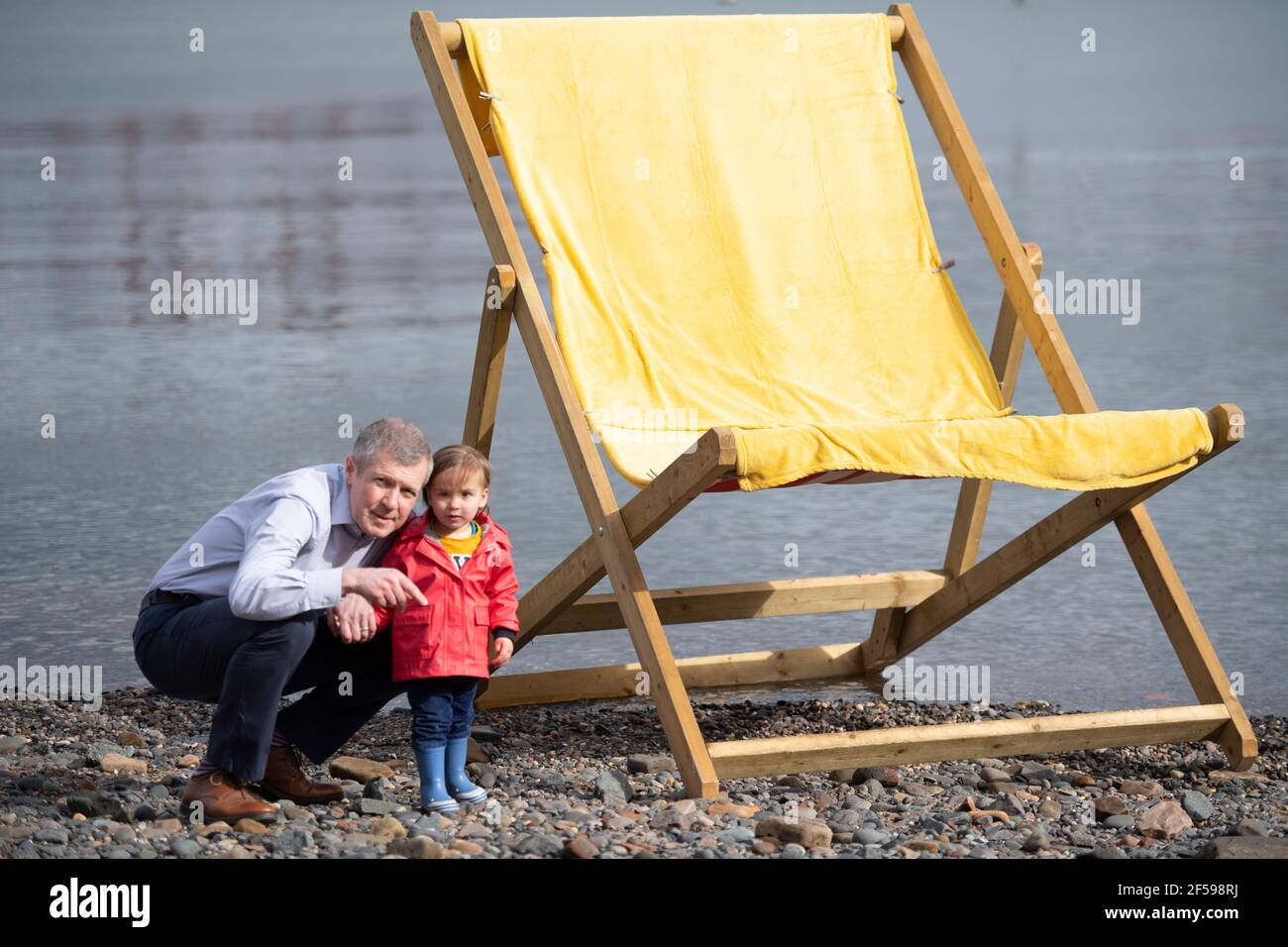 South Queensferry, Scozia, Regno Unito. 25 Marzo 2021. NELLA FOTO: Willie Rennie MSP con Daphne che è la figlia di Rebecca Bell, candidato. Willie Rennie MSP - leader del Partito Liberale democratico Scozzese (Scottish Lib Dems) si è Unito a Rebecca Bell, candidato di Edimburgo Northern e Leith, e a sua figlia Daphne. Legge la figlia di Rebecca un libro su una sedia gigante da spiaggia con la vista del Forth Bridge dietro come parte del loro percorso di campagna elettorale Holyrood per il 6 maggio. Credit: Colin Fisher/Alamy Live News Foto Stock