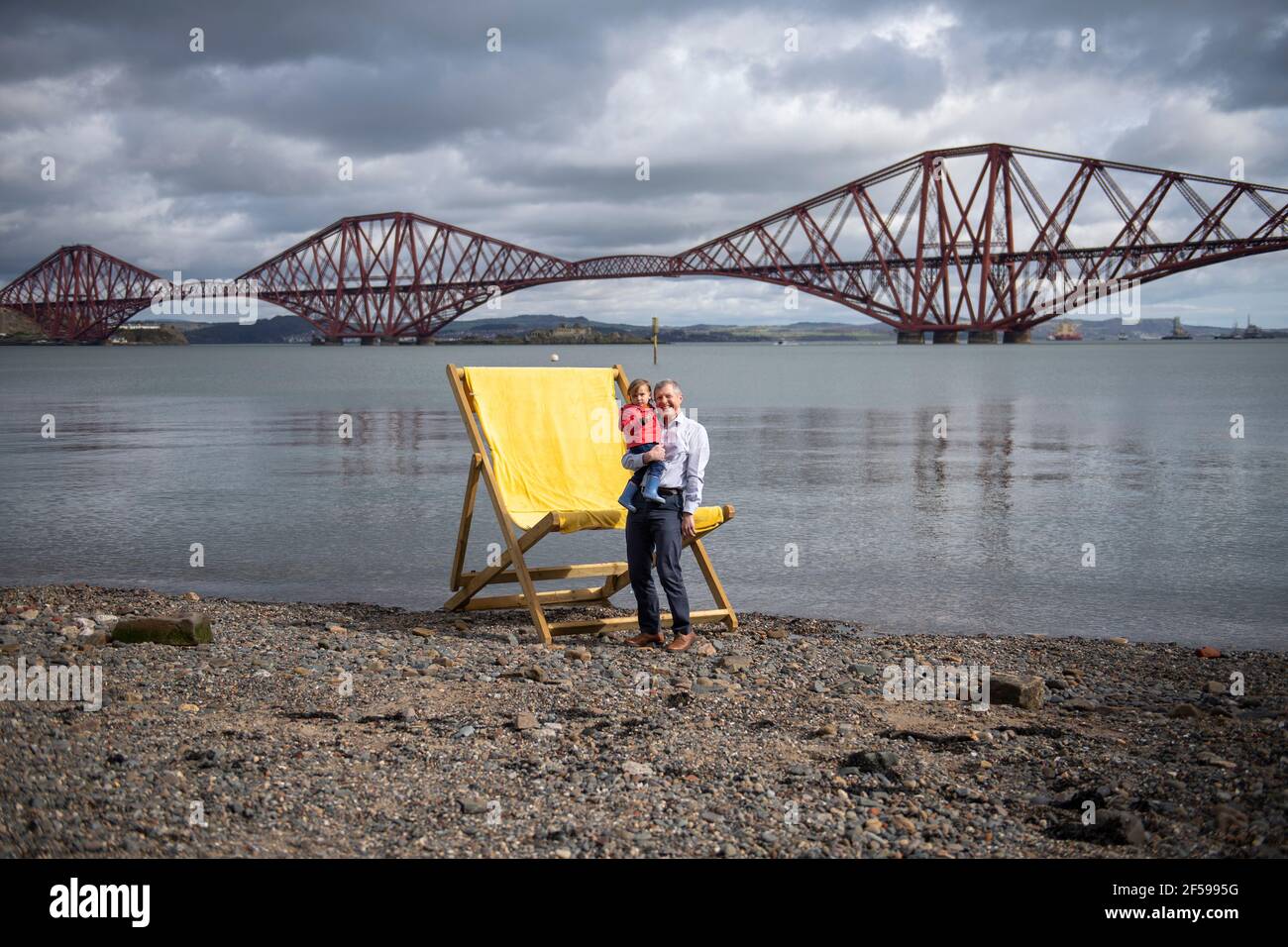 South Queensferry, Scozia, Regno Unito. 25 Marzo 2021. NELLA FOTO: Willie Rennie MSP con Daphne che è la figlia di Rebecca Bell, candidato. Willie Rennie MSP - leader del Partito Liberale democratico Scozzese (Scottish Lib Dems) si è Unito a Rebecca Bell, candidato di Edimburgo Northern e Leith, e a sua figlia Daphne. Legge la figlia di Rebecca un libro su una sedia gigante da spiaggia con la vista del Forth Bridge dietro come parte del loro percorso di campagna elettorale Holyrood per il 6 maggio. Credit: Colin Fisher/Alamy Live News Foto Stock