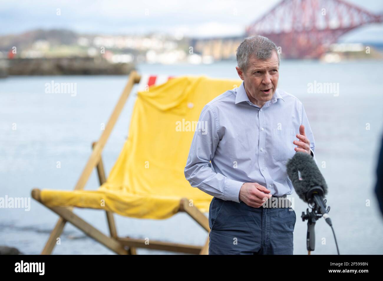 South Queensferry, Scozia, Regno Unito. 25 Marzo 2021. NELLA FOTO: Willie Rennie MSP. Willie Rennie MSP - leader del Partito Liberale democratico Scozzese (Scottish Lib Dems) si è Unito a Rebecca Bell, candidato di Edimburgo Northern e Leith, e a sua figlia Daphne. Legge la figlia di Rebecca un libro su una sedia gigante da spiaggia con la vista del Forth Bridge dietro come parte del loro percorso di campagna elettorale Holyrood per il 6 maggio. Credit: Colin Fisher/Alamy Live News Foto Stock