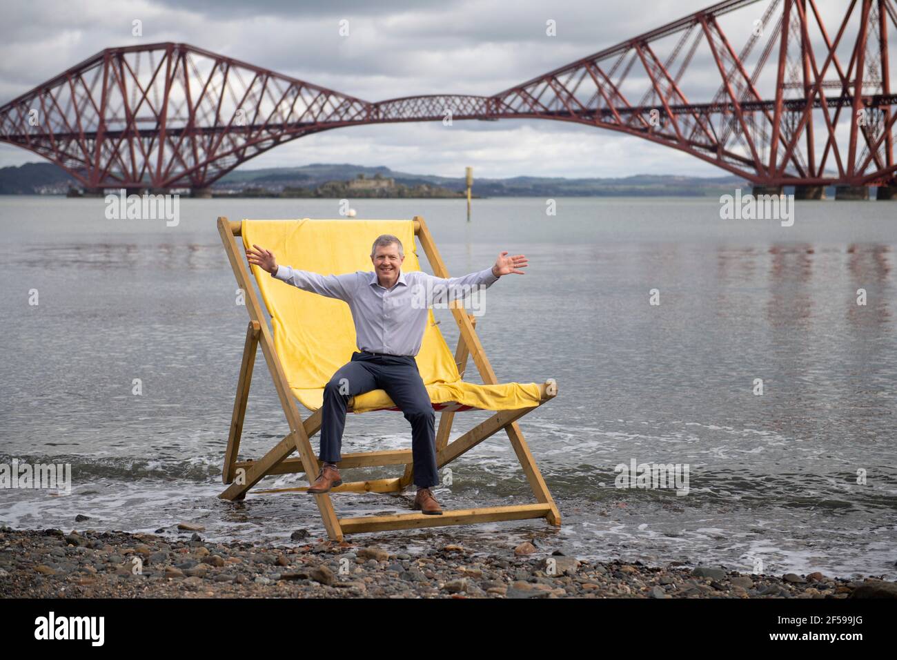 South Queensferry, Scozia, Regno Unito. 25 Marzo 2021. NELLA FOTO: Willie Rennie MSP. Willie Rennie MSP - leader del Partito Liberale democratico Scozzese (Scottish Lib Dems) si è Unito a Rebecca Bell, candidato di Edimburgo Northern e Leith, e a sua figlia Daphne. Legge la figlia di Rebecca un libro su una sedia gigante da spiaggia con la vista del Forth Bridge dietro come parte del loro percorso di campagna elettorale Holyrood per il 6 maggio. Credit: Colin Fisher/Alamy Live News Foto Stock