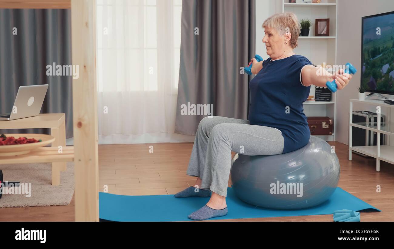Nonna sollevamento pesi seduta sulla palla di bilanciamento in soggiorno. Allenamento di anziani a casa sport stile di vita sano, allenamento di esercizi di fitness anziani in appartamento, attività e assistenza sanitaria Foto Stock