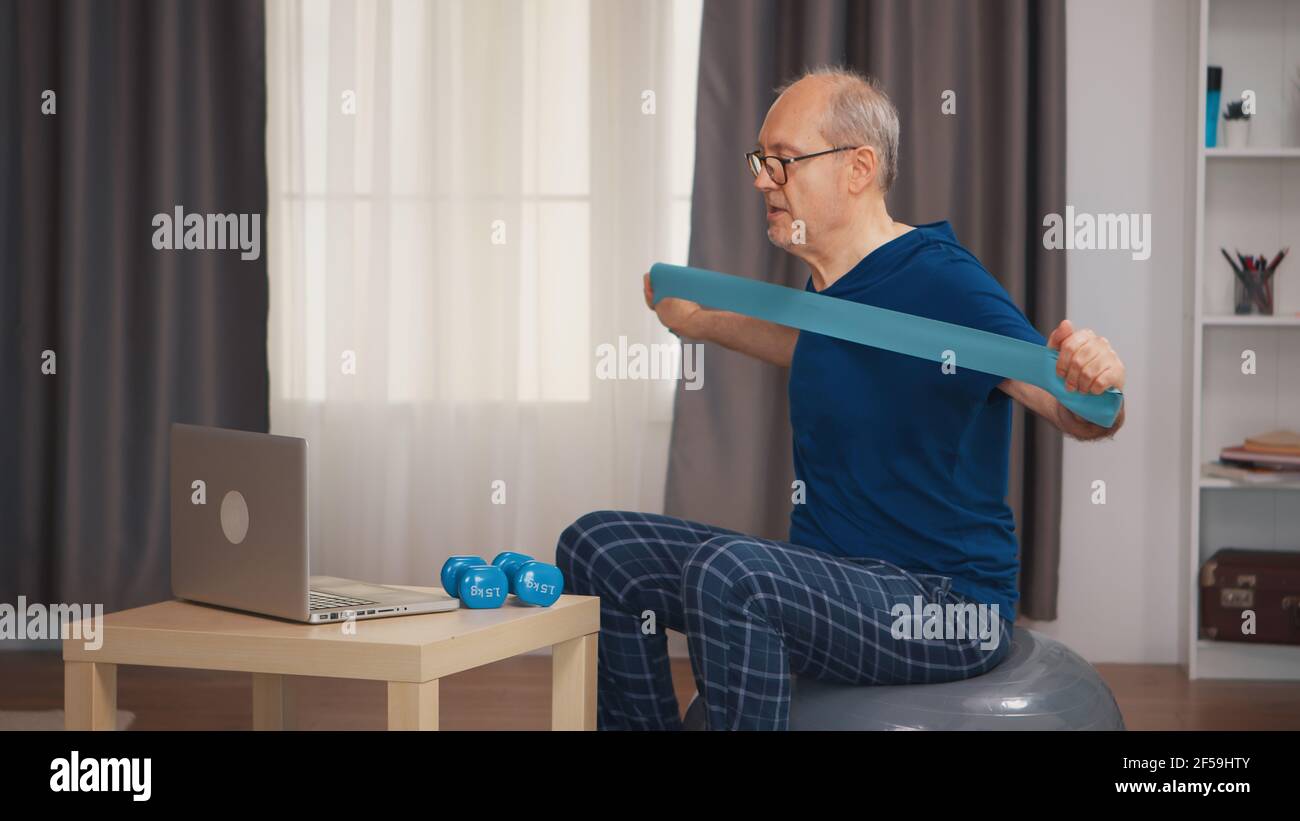 Uomo anziano con vitalità fare esercizio di fitness con banda di resistenza guardando programma online. Anziano pensionato sano addestrando sport di salute a casa, esercitandosi attività di idoneità all'età anziana Foto Stock