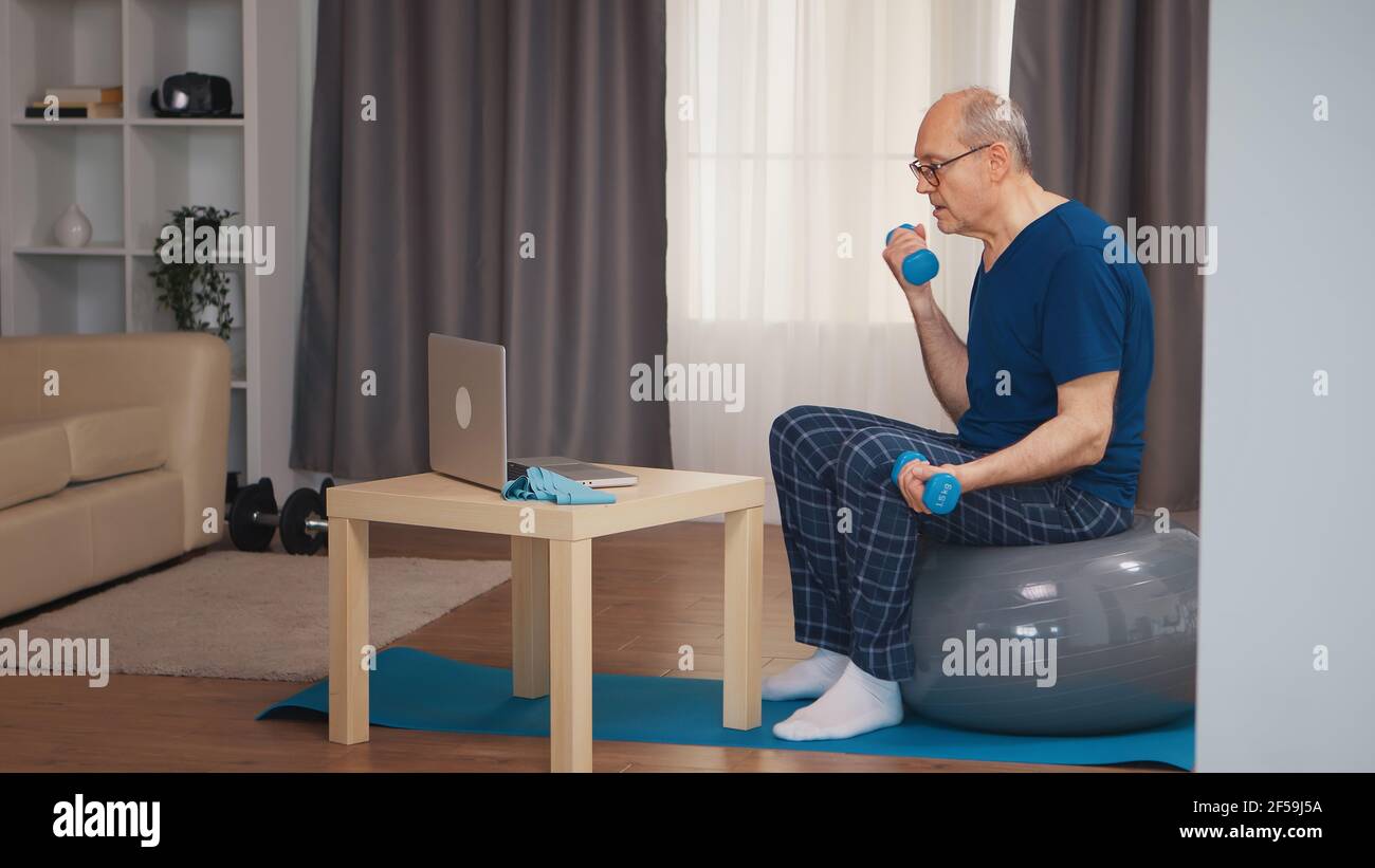 Allenamento uomo anziano in soggiorno con manubri durante il programma di fitness online. Anziano pensionato sano addestrando sport di salute a casa, esercitandosi attività di idoneità all'età anziana Foto Stock