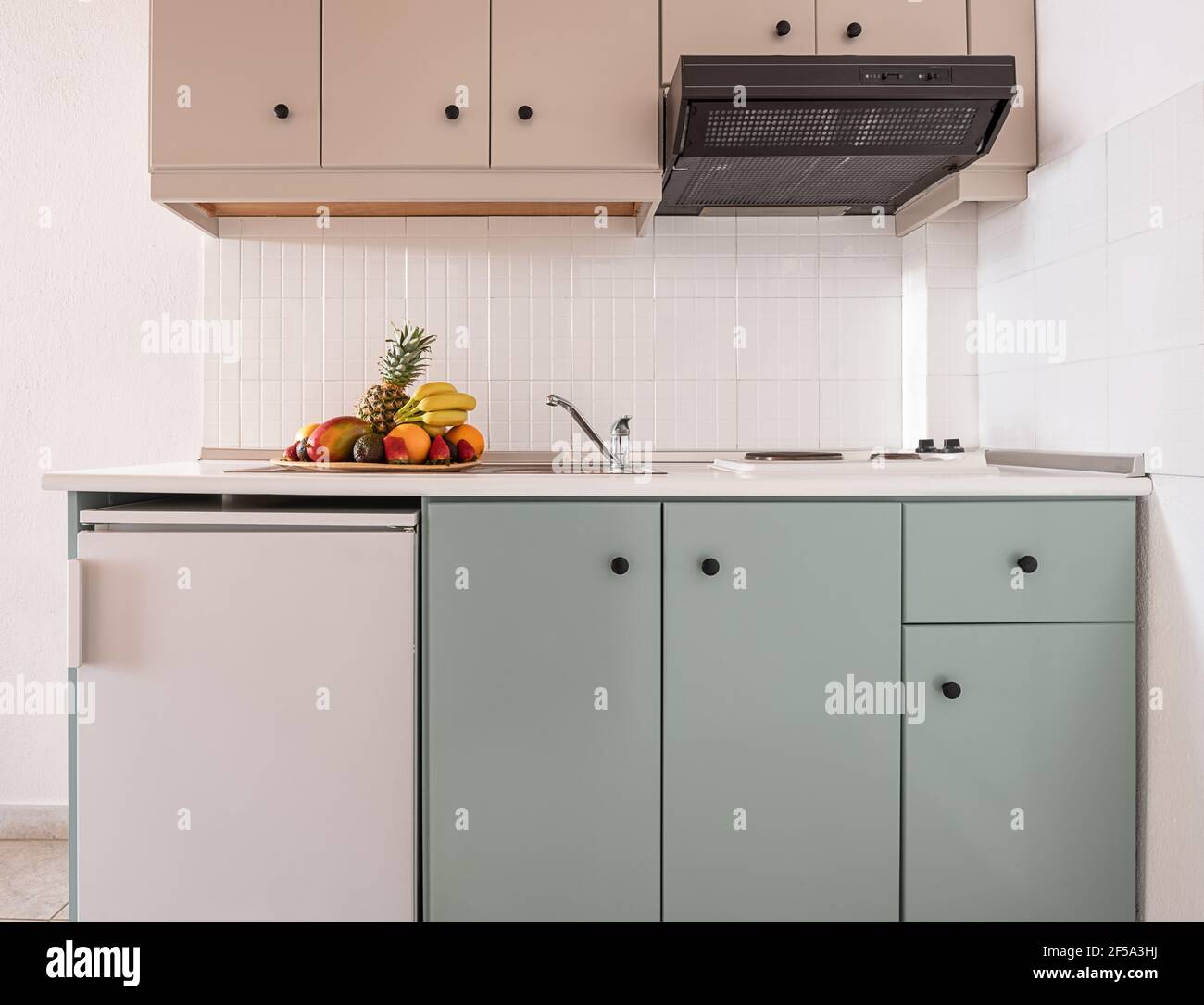 Closeup di piastrelle bianche splashback su parete vuota in semplice armadio modulo di moderna cucina interna con piccolo frigorifero, sfiato, lavello, piano cottura, piatto di frutta Foto Stock