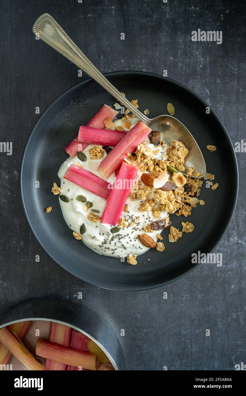 Piatto di yogurt e rabarbaro in camicia con granola in una ciotola nera e una padella con rabarbaro su fondo nero, con spazio copia, verticale Foto Stock