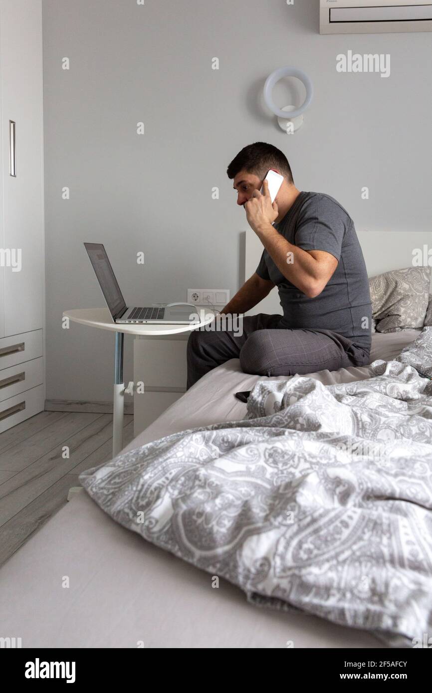 Uomo che parla al telefono, lavorando dietro un computer portatile in camera da letto sul Foto Stock