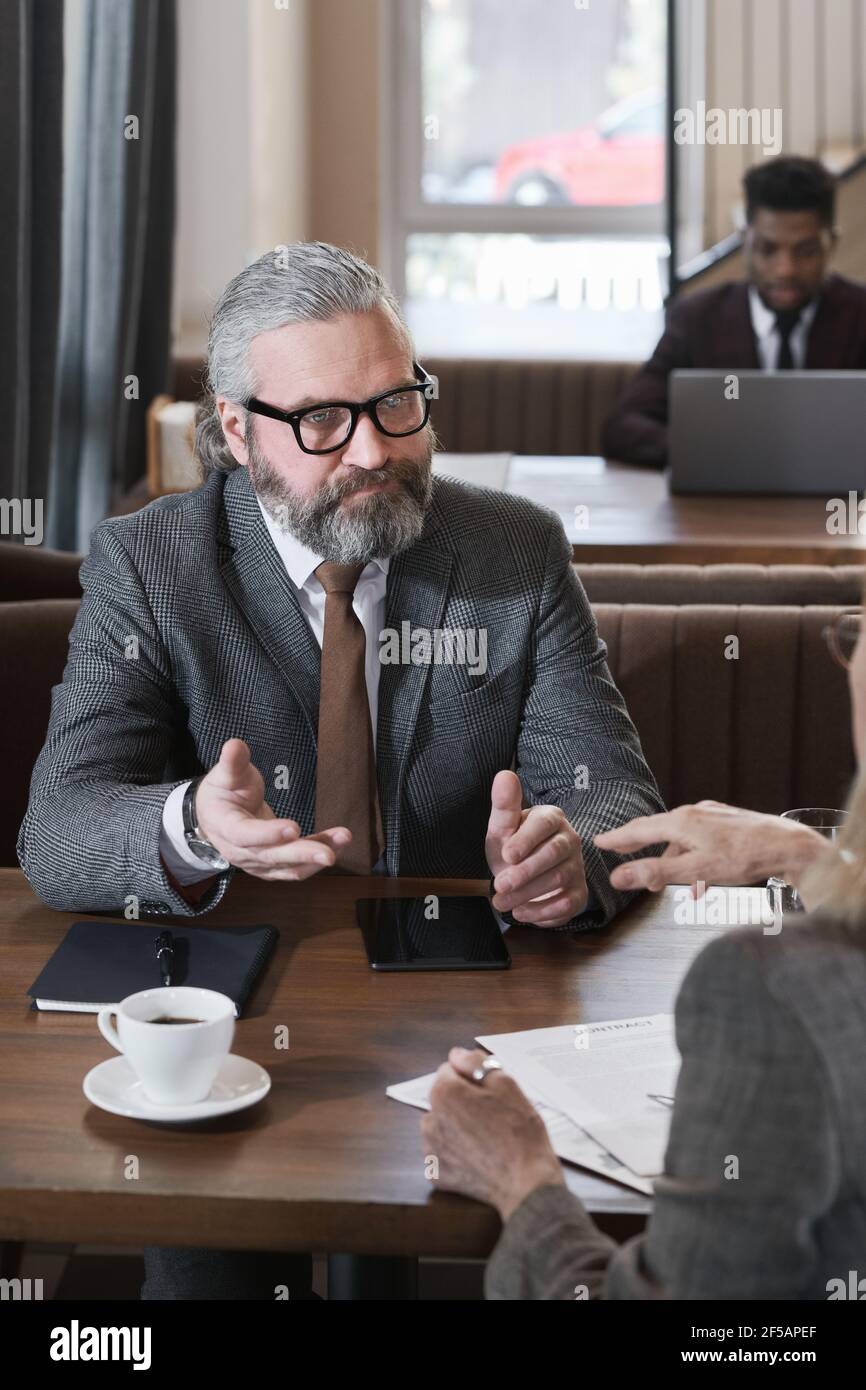 Uomo d'affari maturo bearded seduto al tavolo con il suo collega e discutere di lavoro durante la riunione nel ristorante Foto Stock