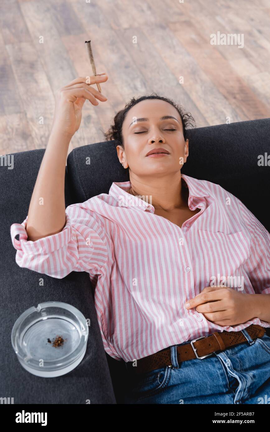 vista ad alto angolo della donna afroamericana sdraiata sul divano e il fumo rotolò unito con cannabis medica vicino al posacenere Foto Stock
