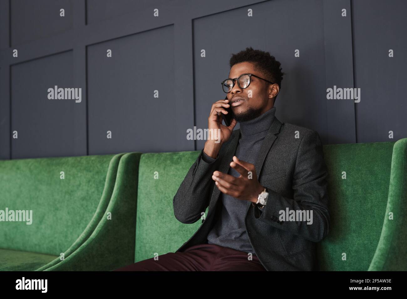 Giovane africano che parla con il suo collega sul telefono cellulare seduta su un bellissimo divano verde Foto Stock