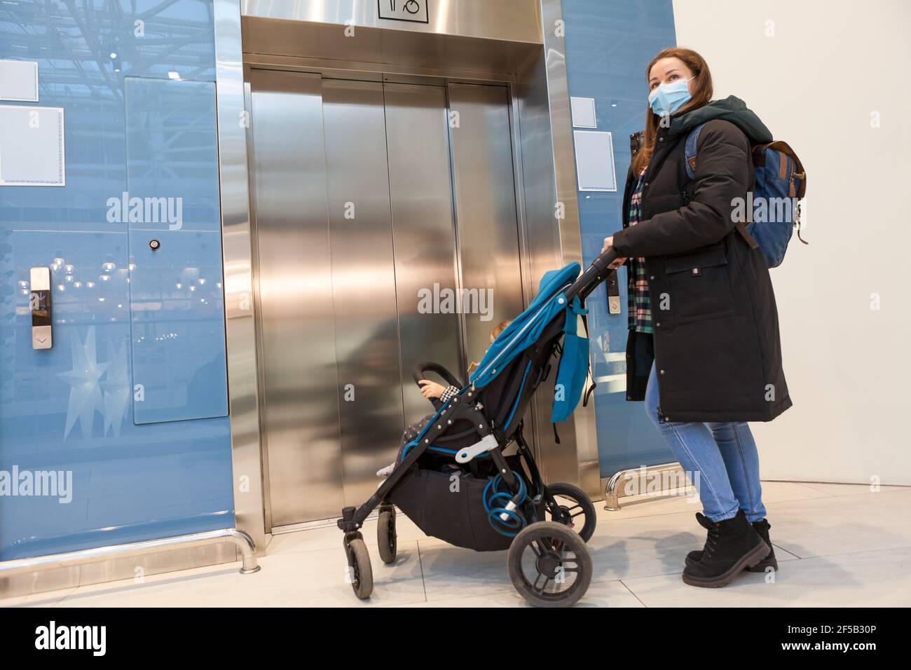 Madre che indossa maschera viso in attesa ascensore con bambini PRAM in hall di un centro commerciale Foto Stock