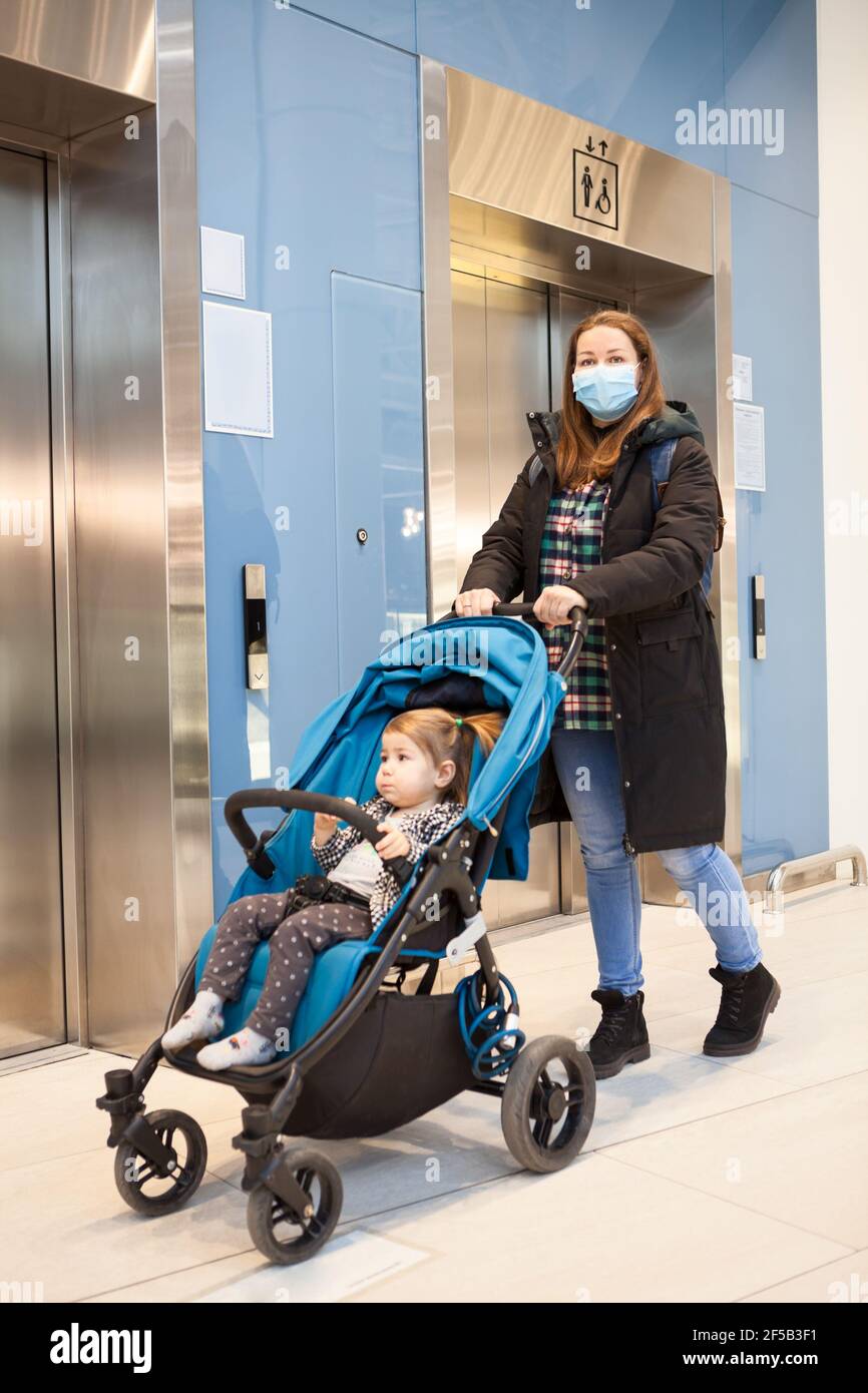 Madre adulta che indossa una maschera medica proveniente dall'ascensore con passeggino per bambini, sala di un centro commerciale Foto Stock