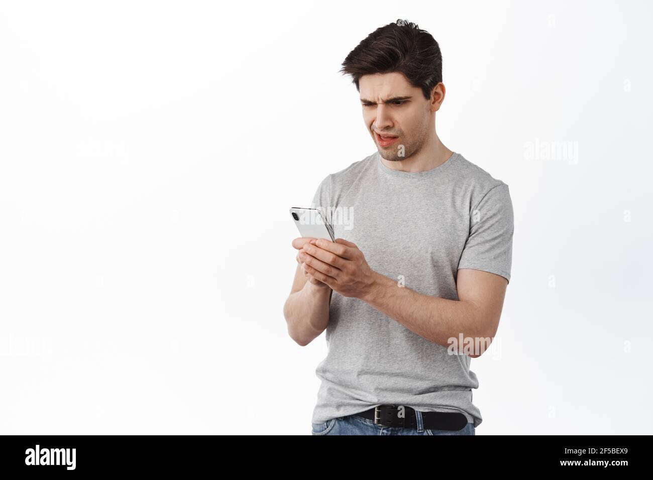 Uomo guardare confuso al telefono cellulare, leggere strano messaggio sullo schermo del cellulare, ricevere cattive notizie su smartphone social media, sfondo bianco Foto Stock