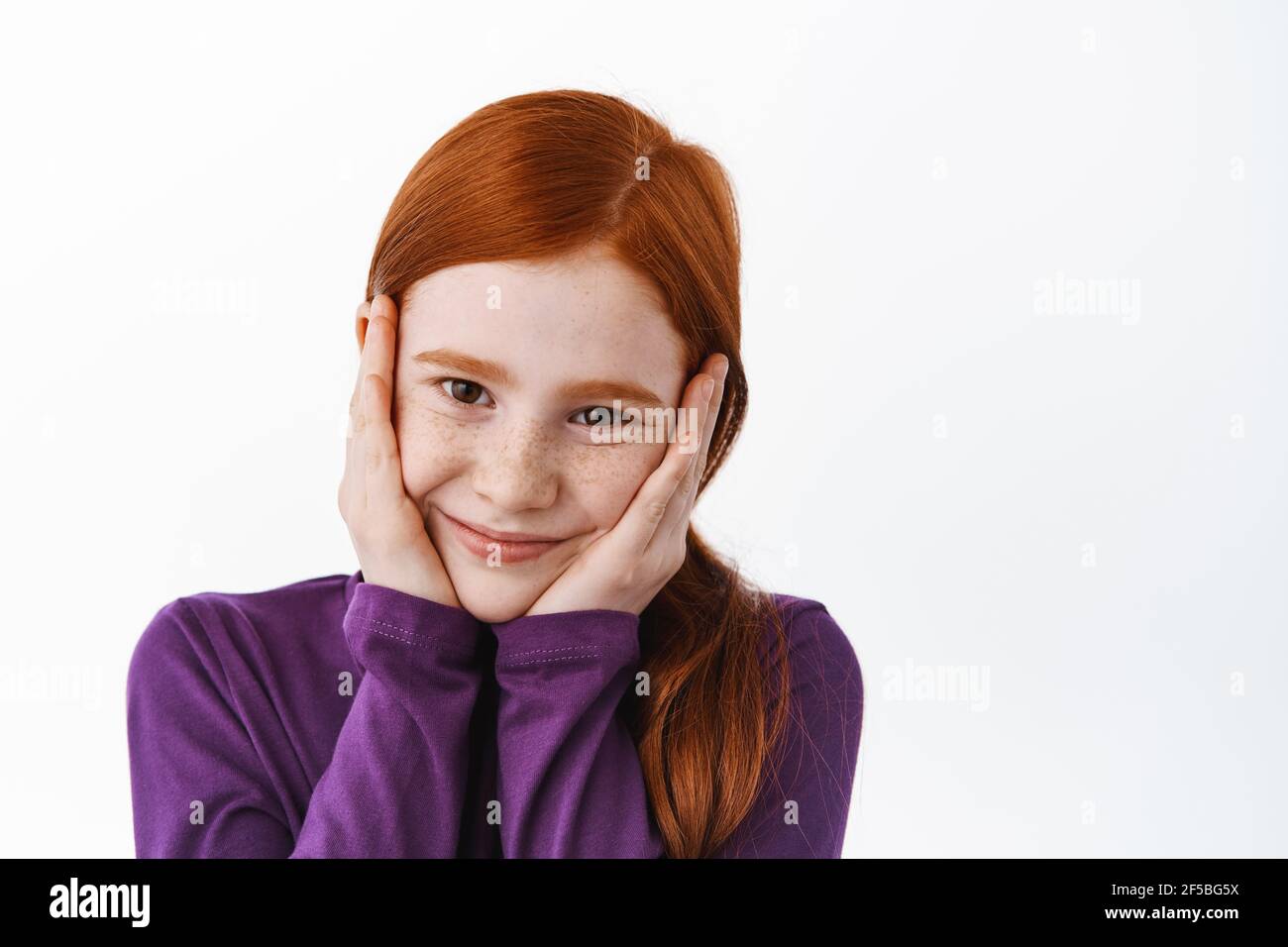 Adorabile bambina con capelli ricci e zenzero, tenendo le mani sulle guance e sorridendo, arrossendo carino, guardando gioioso alla macchina fotografica, sfondo bianco Foto Stock