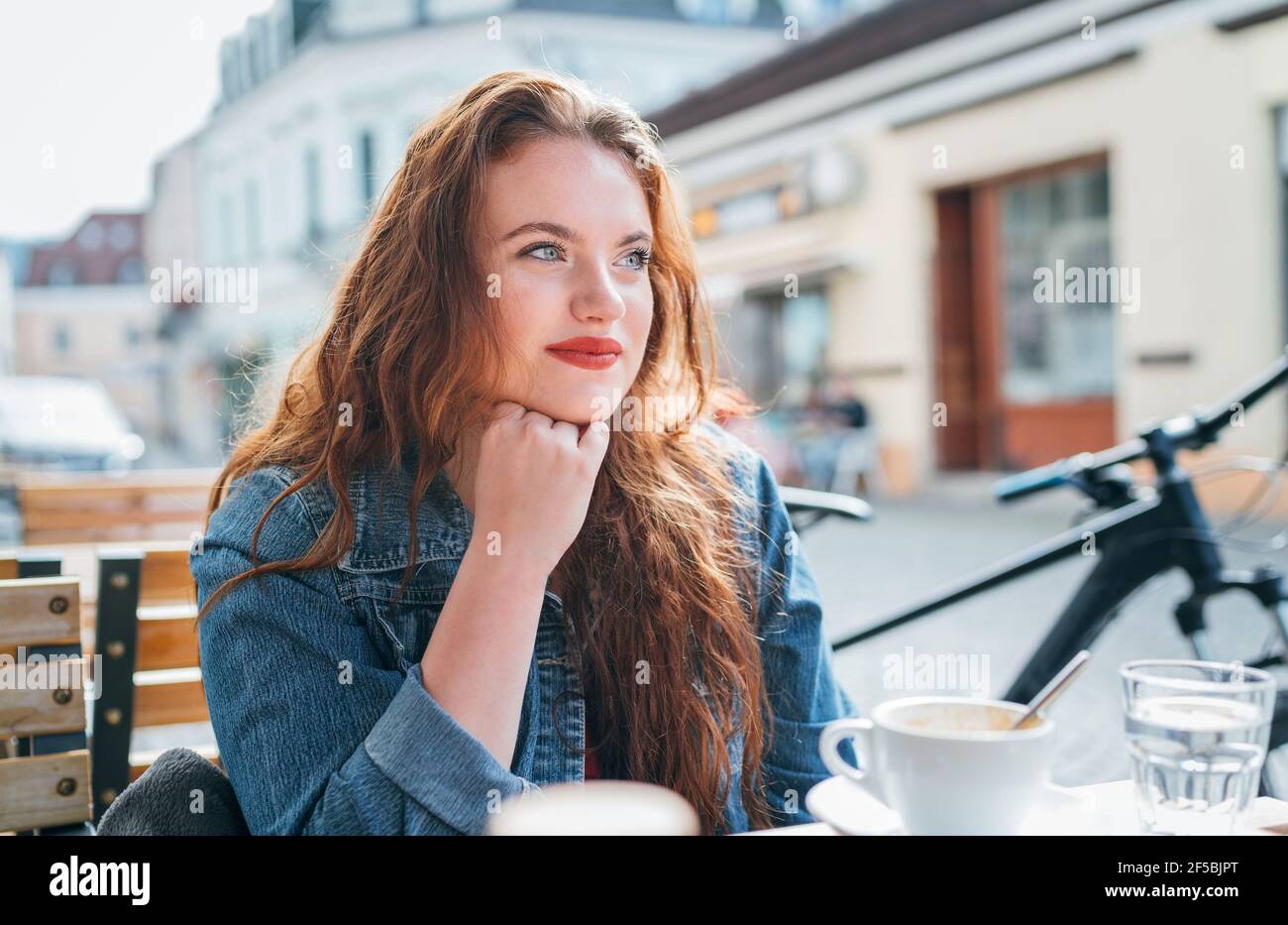 Ritratto di triste rosso arricciato capelli lunghi caucasica donna seduta su un accogliente caffè all'aperto terrazza sulla strada e guardando la strada. Giovane donna takin Foto Stock