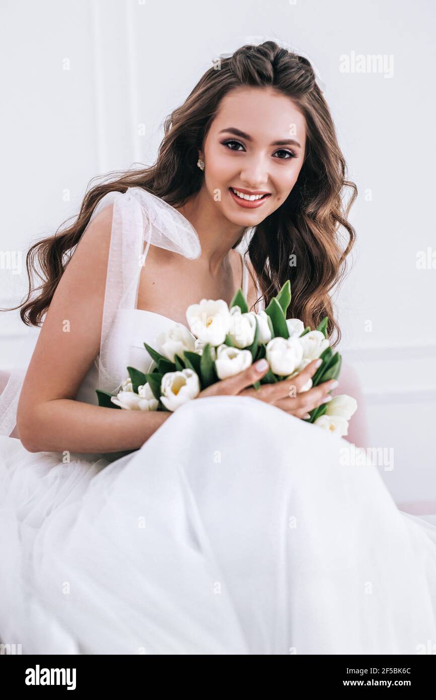 Sposa in bellissimo abito tenuta bouquet di nozze e seduta su divano interno Foto Stock