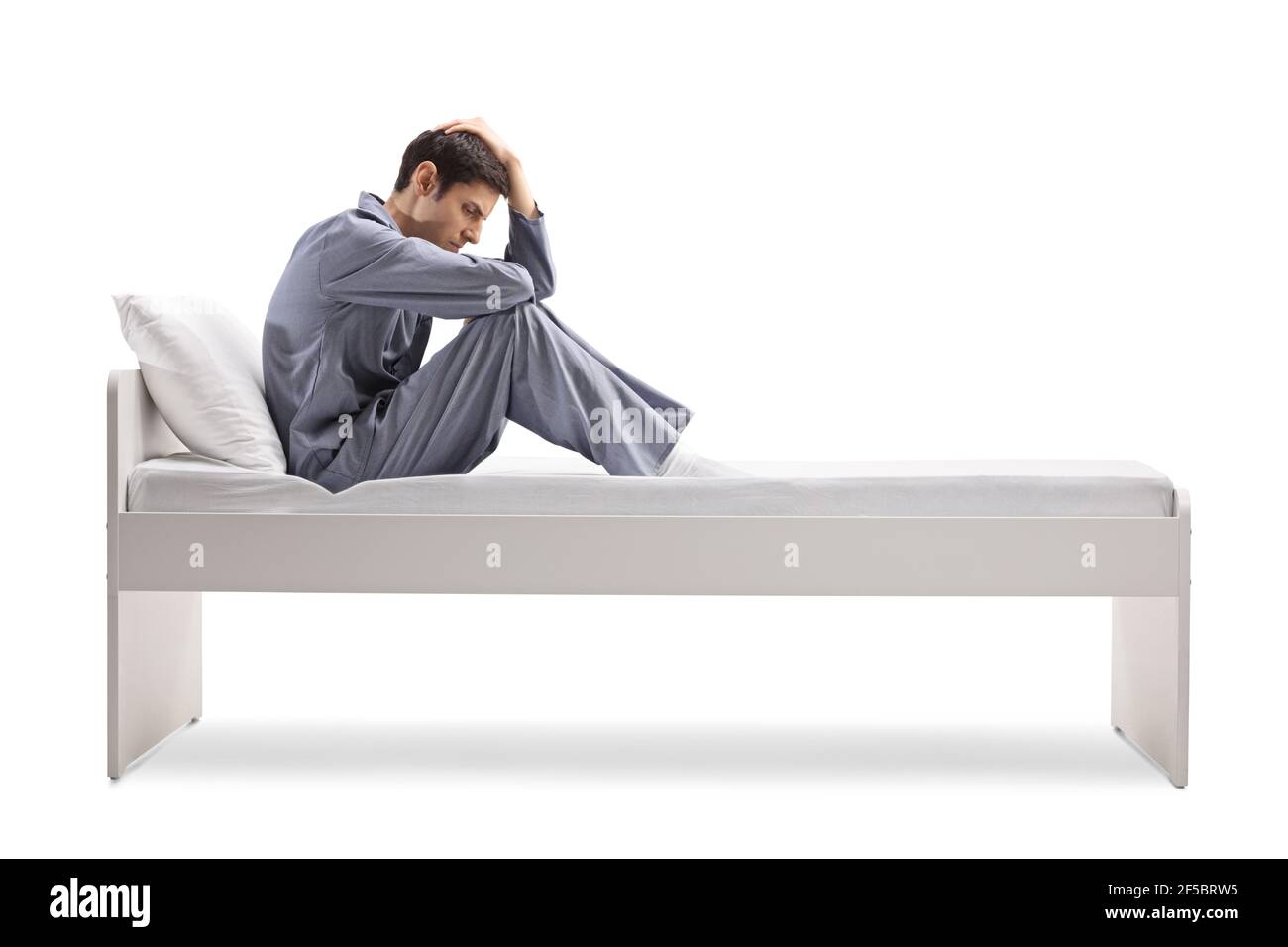 Foto di un uomo triste in pigiama a tutta lunghezza seduta su un letto singolo isolato su sfondo bianco Foto Stock