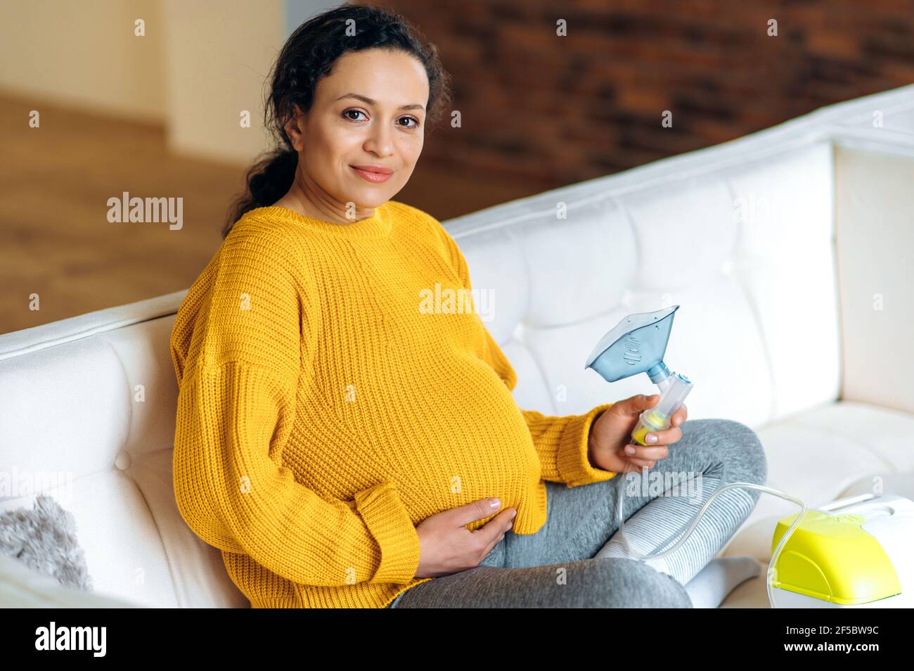 Malata donna incinta esausta razza mista, seduta sul divano nel soggiorno, indossando un maglione, sottoponendosi a trattamento di inalazione, tenendo l'inalatore in mano, guardando la macchina fotografica Foto Stock