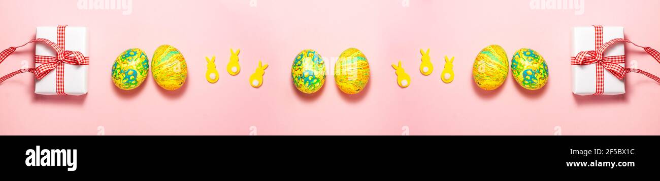 Striscione di uova di Pasqua verdi e gialle, scatola regalo bianca con prua regalo rossa e conigli di velluto giallo con code bianche su sfondo rosa brillante. Piatto la Foto Stock