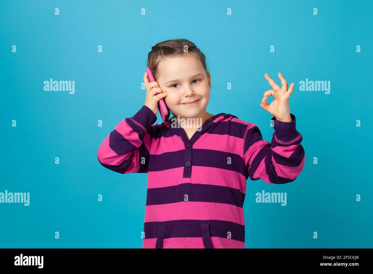 Ritratto di una ragazza carina e allegra in un viola striato parlando su un telefono cellulare e mostrando OK isolato su uno sfondo blu Foto Stock