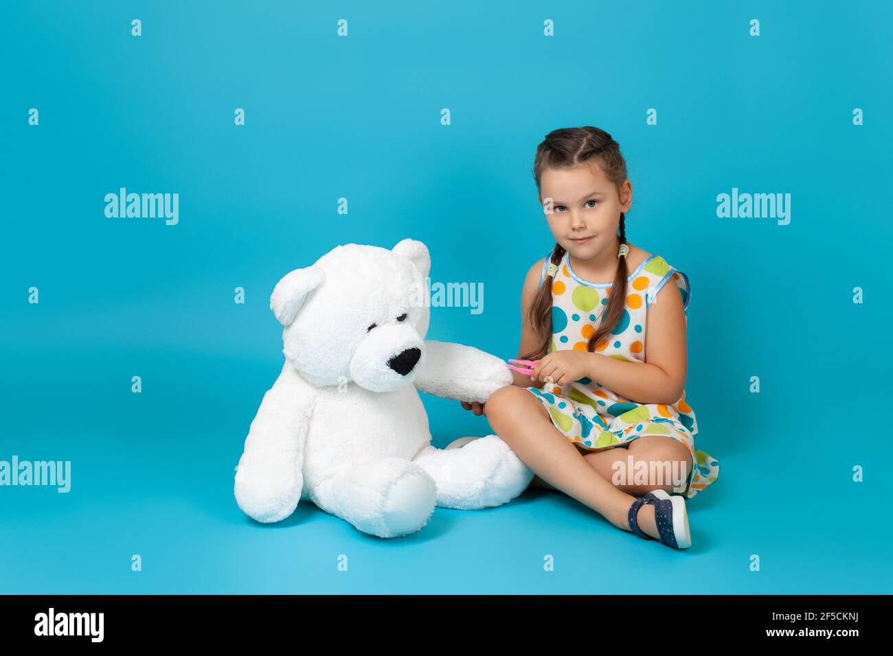 l ragazza seduta a gambe incrociate sul pavimento e tirando un splinter dalla zampa di un orsacchiotto bianco con pinzette isolate su sfondo blu Foto Stock