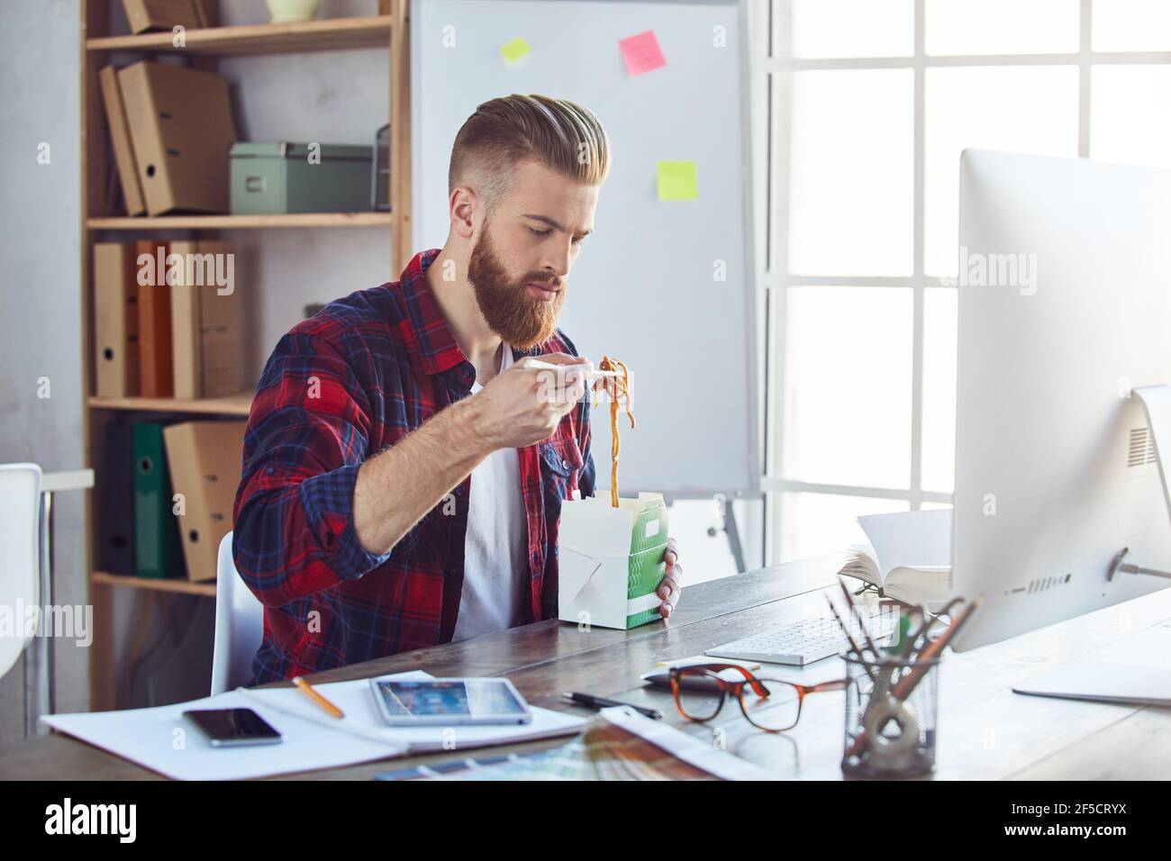 Ragazzo di stile in abbigliamento casual che assaggia cibo asiatico al suo luogo di lavoro Foto Stock