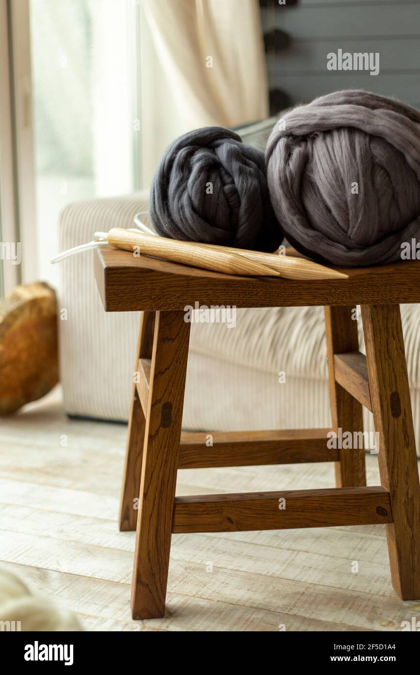 Due grandi palle di lana merino adagiate su un legno sgabello con aghi in legno da maglieria sullo sfondo di a. finestra e divano bianco Foto Stock