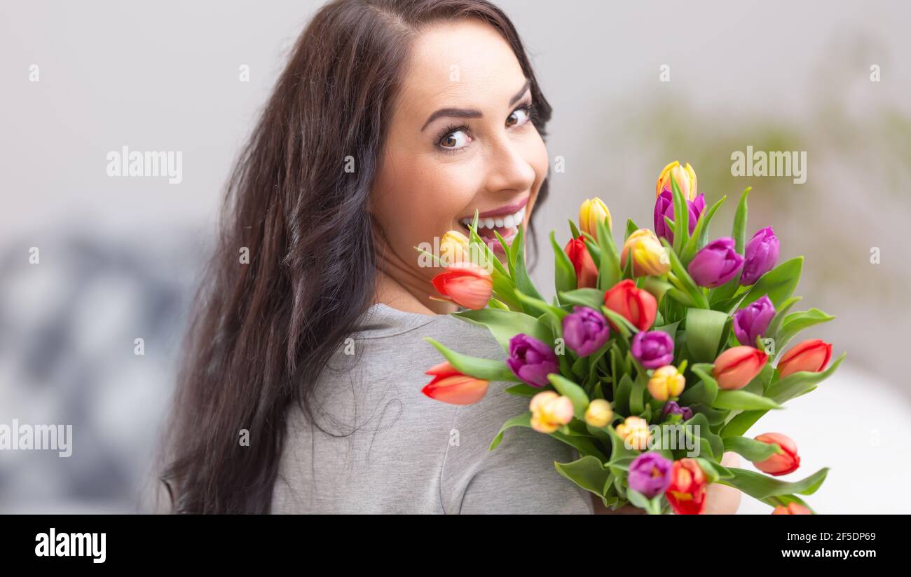 Felice donna dai capelli scuri che sorride e tiene un bel bouquet pieno di tulipani durante la giornata nazionale delle donne. Foto Stock