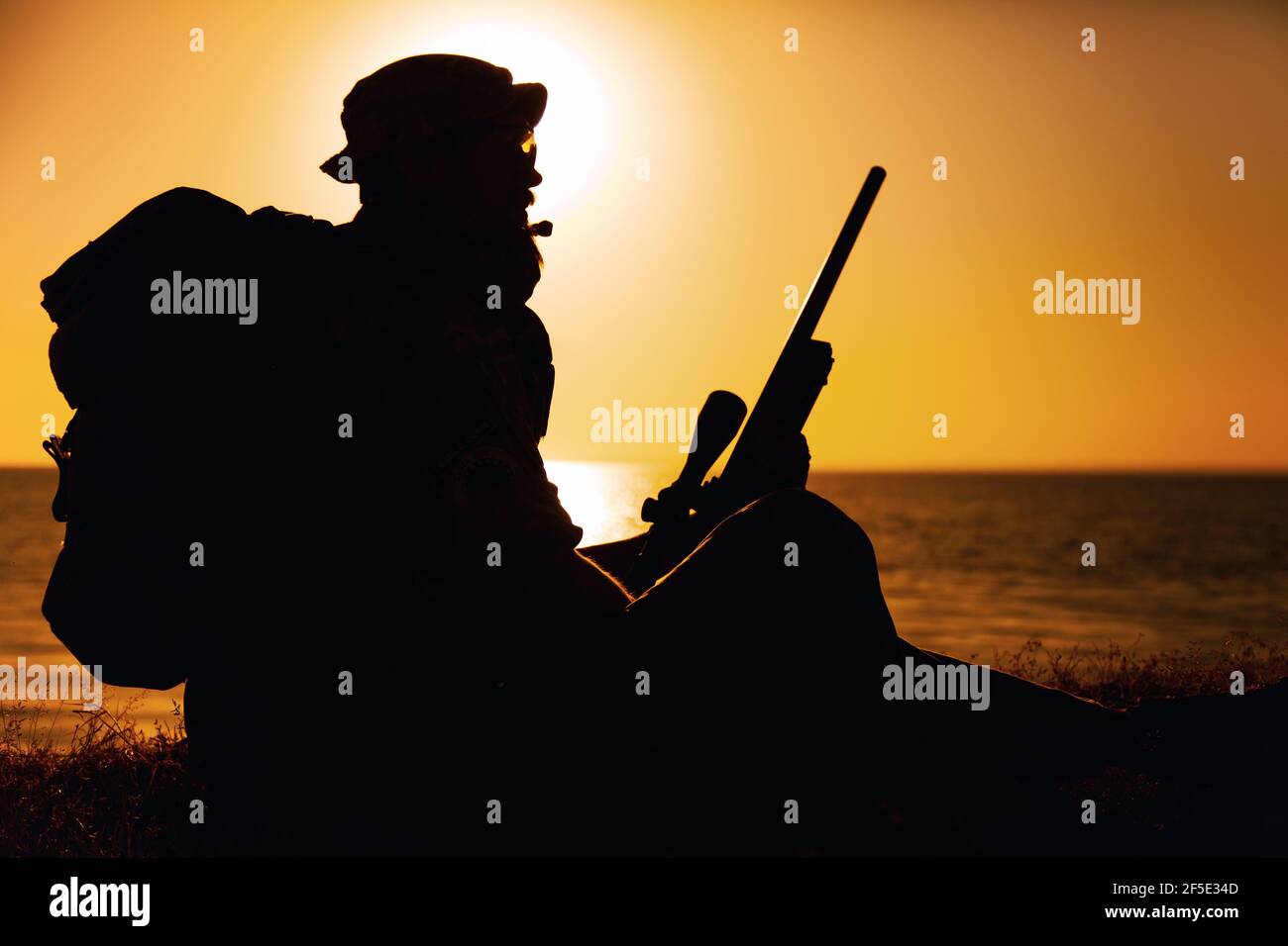 Silhouette di combattente commando, sniper dell'esercito delle forze speciali seduto sul mare o sulla riva dell'oceano al tramonto. Guardia costiera rifleman osservando la spiaggia, riposando durante la pattuglia costiera o operazione anfibia Foto Stock