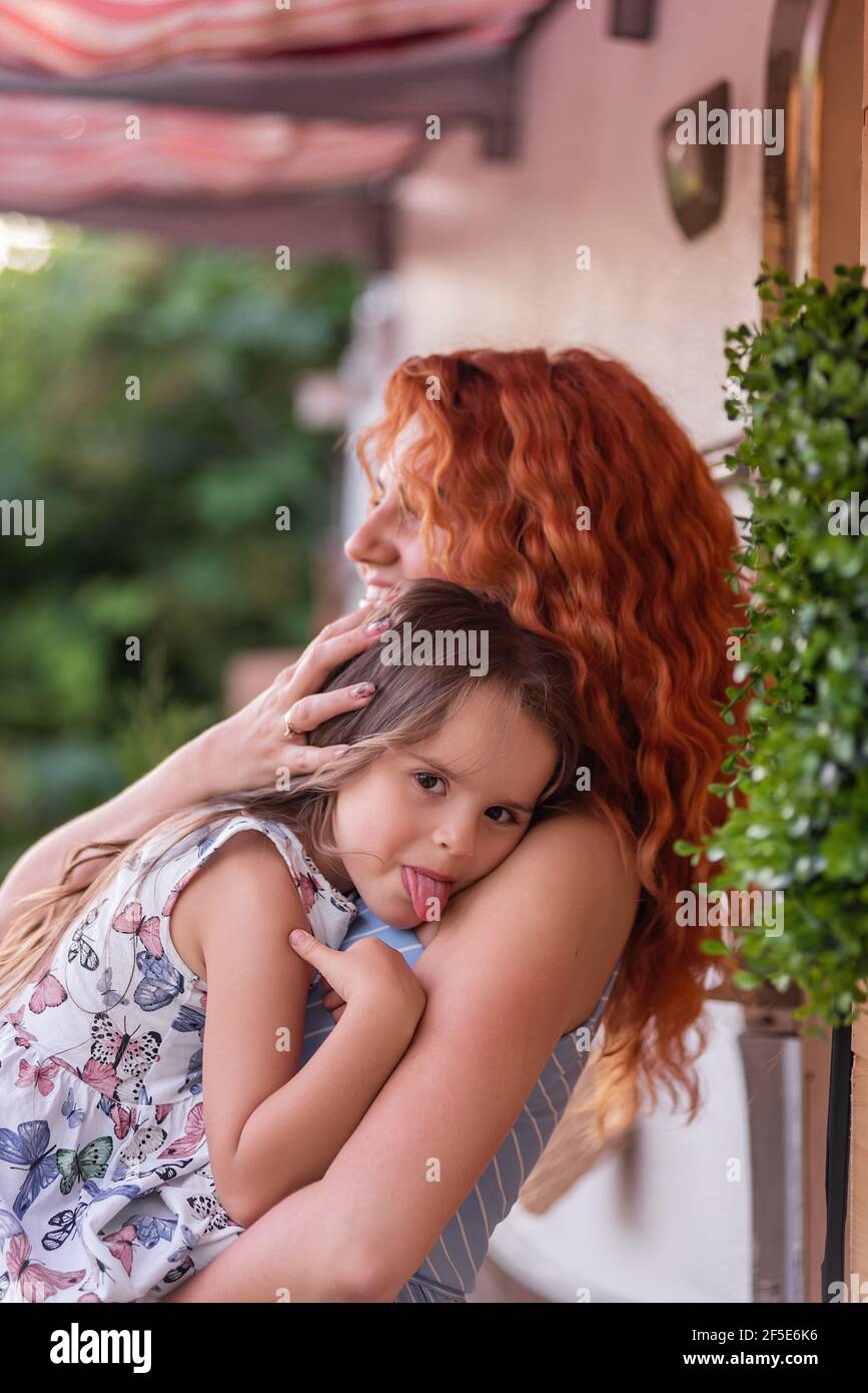 Una bella giovane madre dai capelli rossi tiene gentilmente, abbraccia piccola figlia in armi vicino a casa con camion rimorchio. La ragazza sorride, mostra la lingua alla camma Foto Stock