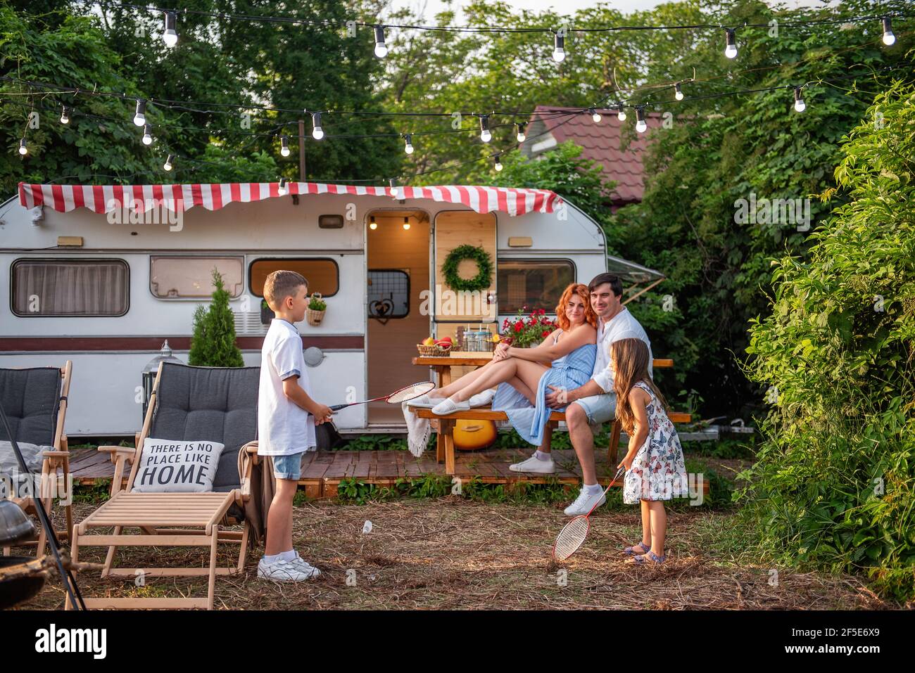 Fratello allegro e sorella che gioca badminton dal camion del rimorchio, genitori felici sono seduti su panca di legno. Padre abbraccia la madre, i bambini hanno divertimento o Foto Stock