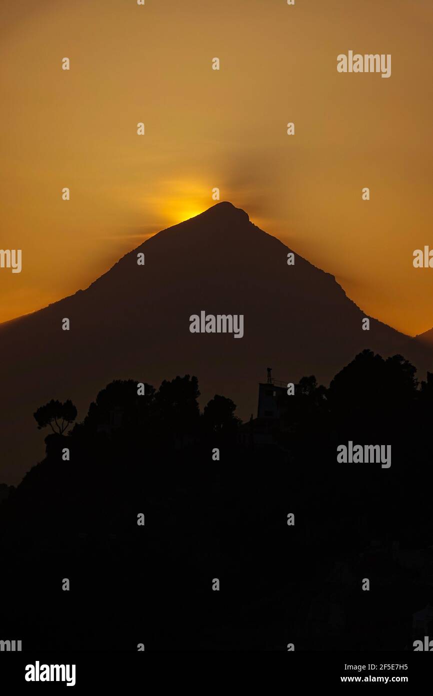 Il sole tramonta dietro la cima triangolare del Puig Campana, vicino a Benidorm, sulla Costa Blanca, in Spagna Foto Stock