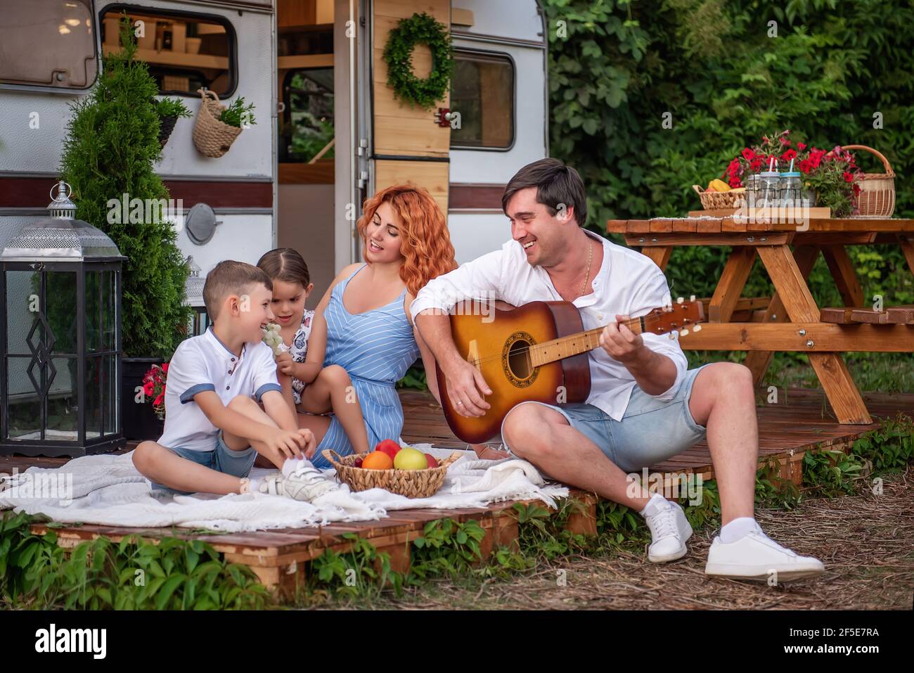 Il giovane padre suona la chitarra, madre dai capelli rossi con bambini che cantano canzoni. Happy Family al pic-nic, campeggio in camion rimorchio nella natura fuori della città Foto Stock