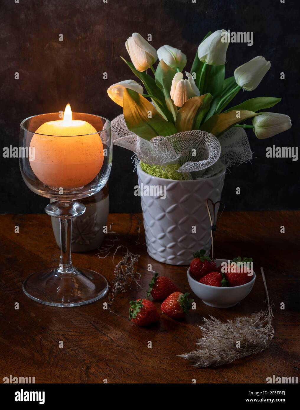 Una candela rotonda è accesa in un portacandele di vetro vicino a un vaso di fiori bianchi su un tavolo di legno con alcune fragole e piante secche Foto Stock