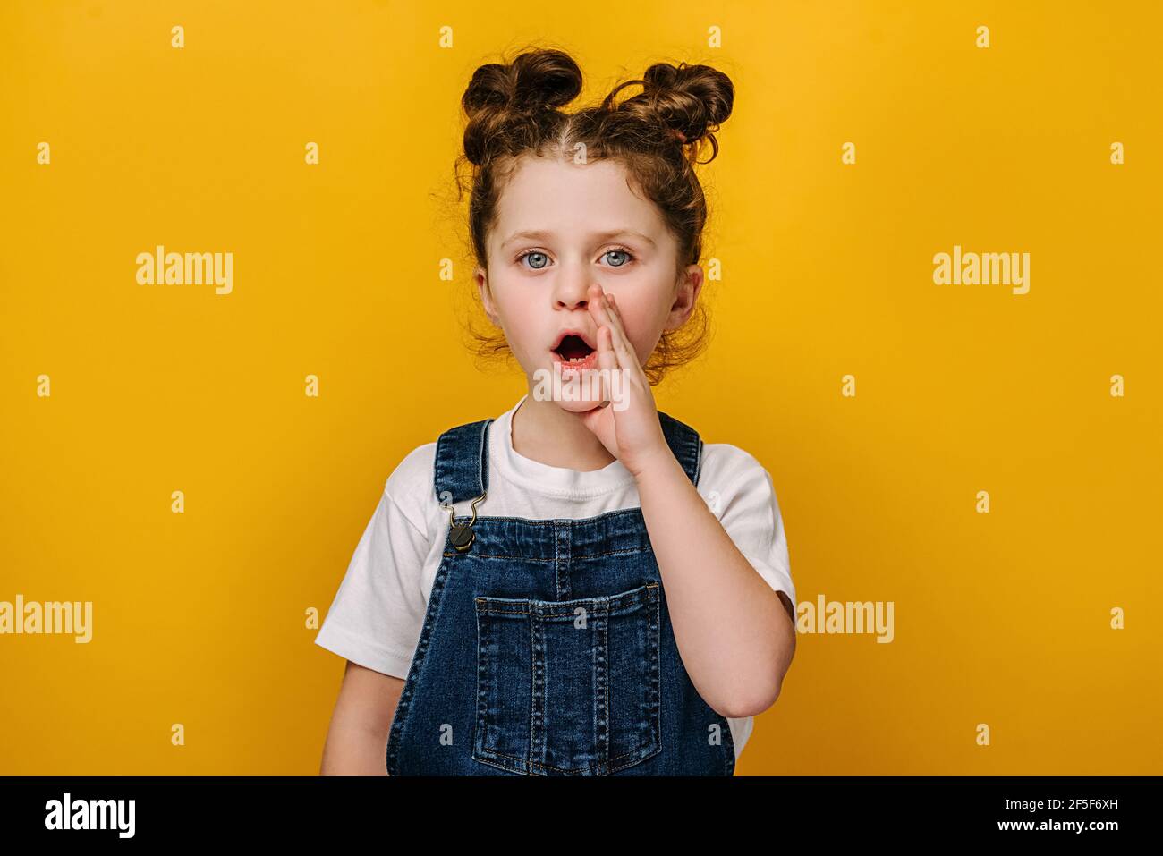 Primo piano ritratto di sorpresa piccola ragazza prescolare tenendo la mano vicino bocca, tranquillamente dicendo segreto, isolato su sfondo giallo studio con copia s. Foto Stock