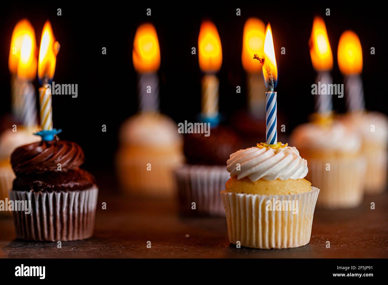 Primo piano immagine vista laterale di un assortimento di cupcake al cioccolato e alla vaniglia con glassature bianche e marroni. Ognuno ha una candela accesa contro bac scuro Foto Stock