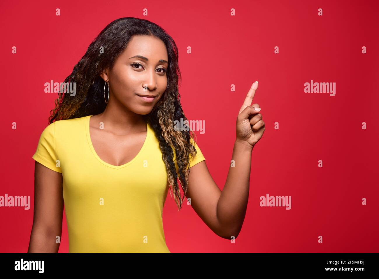 Giovane affascinante ragazza dalla pelle scura in una T-shirt gialla sorride e punta il dito indice verso lo spazio sulla destra. Modello su sfondo rosso brillante cl Foto Stock