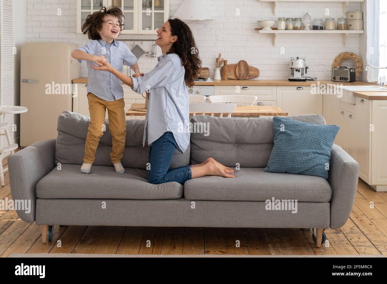 Madre e bambino che ride divertendosi, tenendo le mani e saltando sul divano a casa. Buona famiglia. Foto Stock