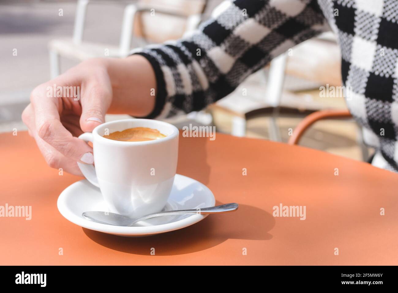 mano di una giovane donna caucasica che tiene una tazza bianca di caffè sulla parte superiore di un tavolo arancione a. una caffetteria con terrazza Foto Stock