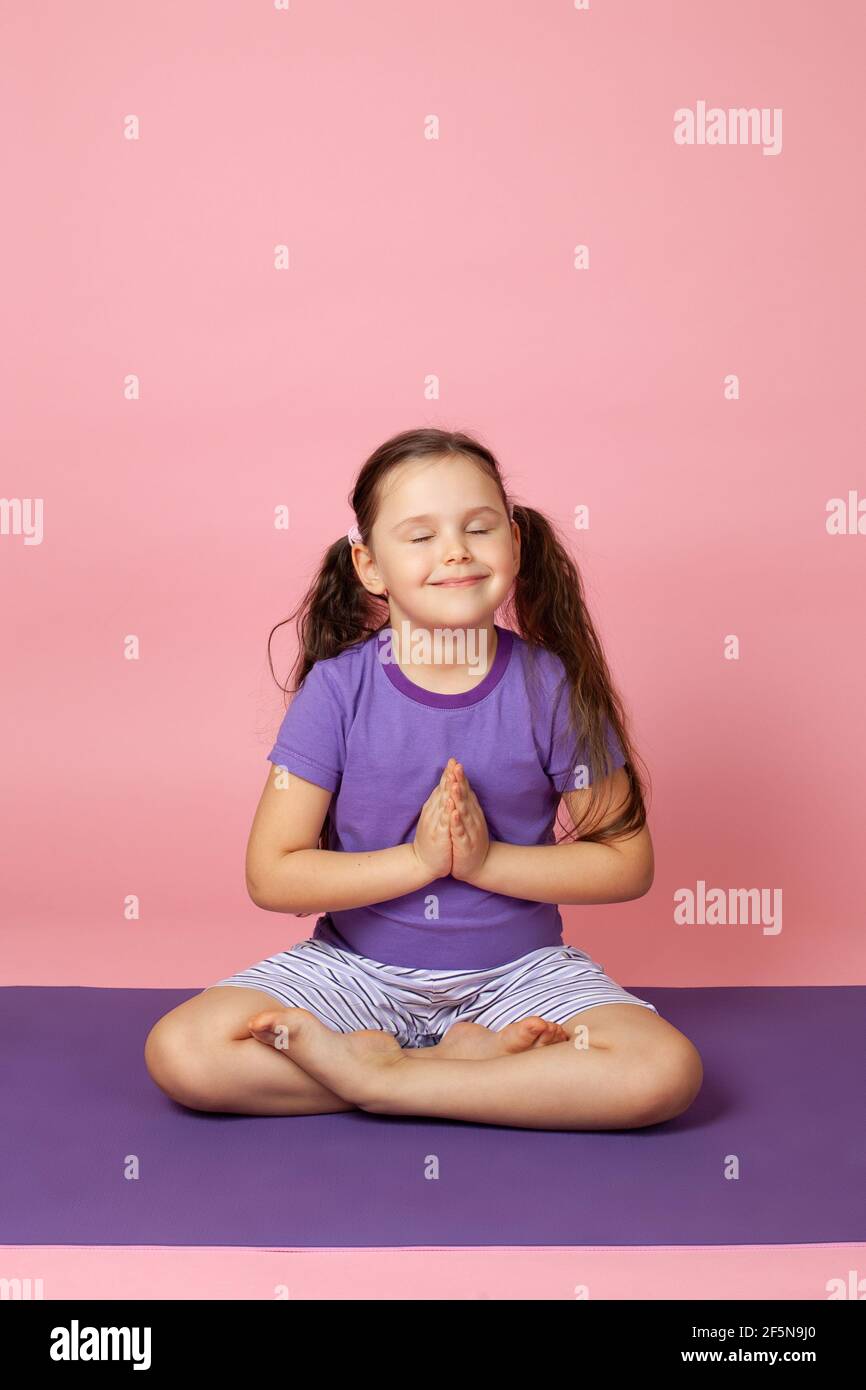 una ragazza felice pacifica di sei anni fa yoga, si siede in una posizione di loto o prega, isolato su uno sfondo rosa Foto Stock