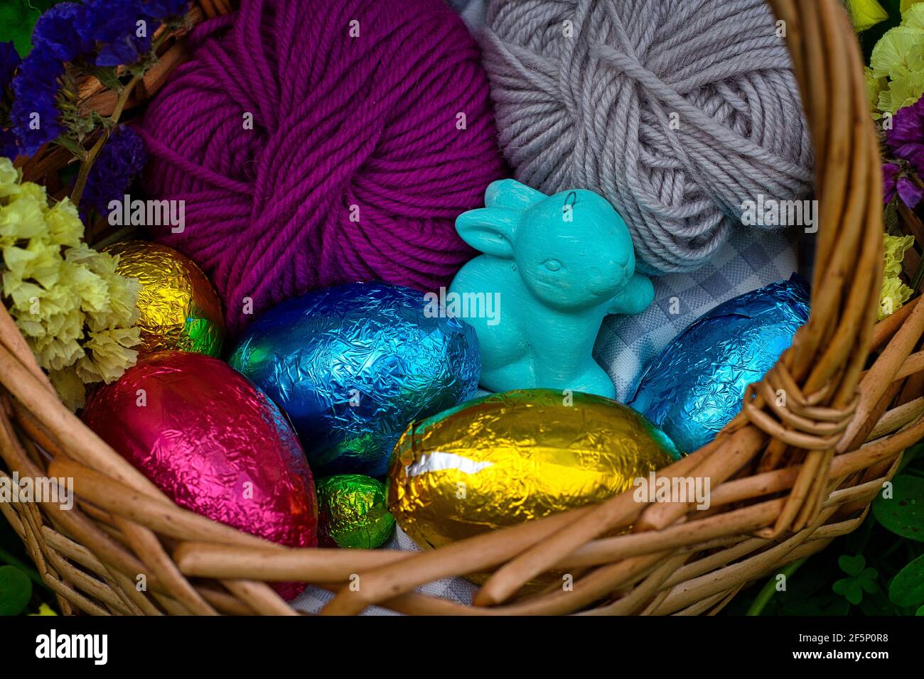 Un cestino decorato per una caccia all'uovo. Ha uova avvolte in foglio di stagno colorato, un giocattolo di un coniglio e alcune palle di filato. E' circondato da vegetazione, cl Foto Stock