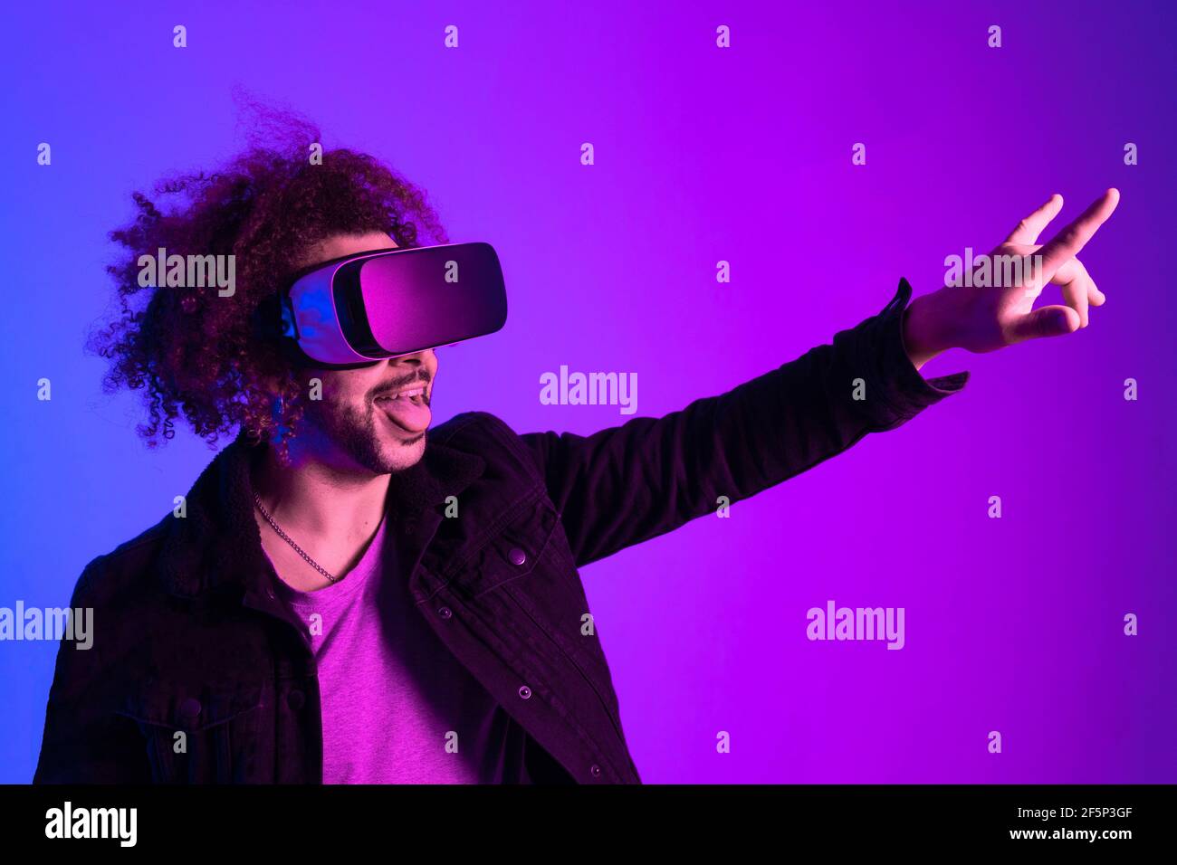 Modello giovane con barba e capelli afro in occhiali di realtà virtuale su lilla e sfondo blu facendo segno di vittoria. Realtà aumentata, tecnologia futura Foto Stock